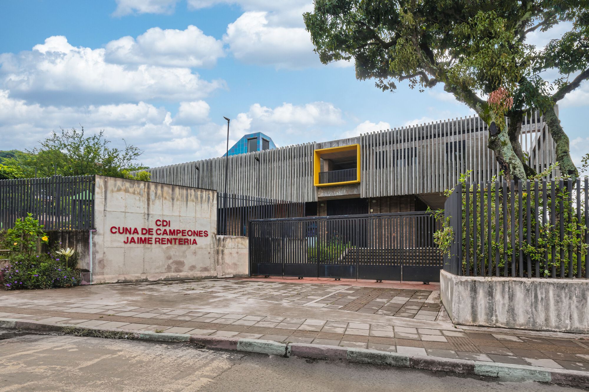 Street view of vertical metal slat facade with yellow window frame and blue pyramid roof beside weathered concrete wall