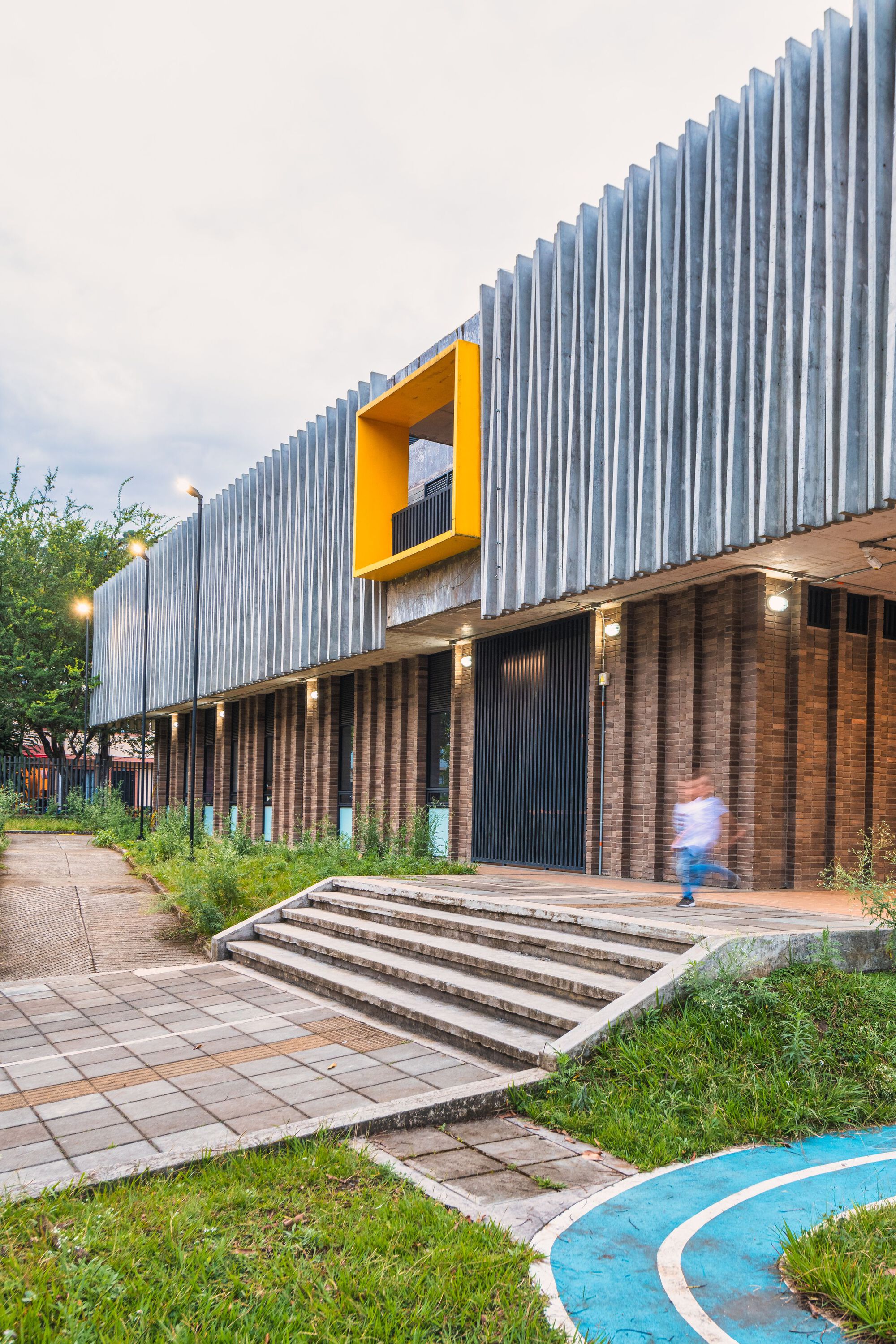 Entrance facade with vertical corrugated metal cladding, yellow window aperture, and brick base at dusk