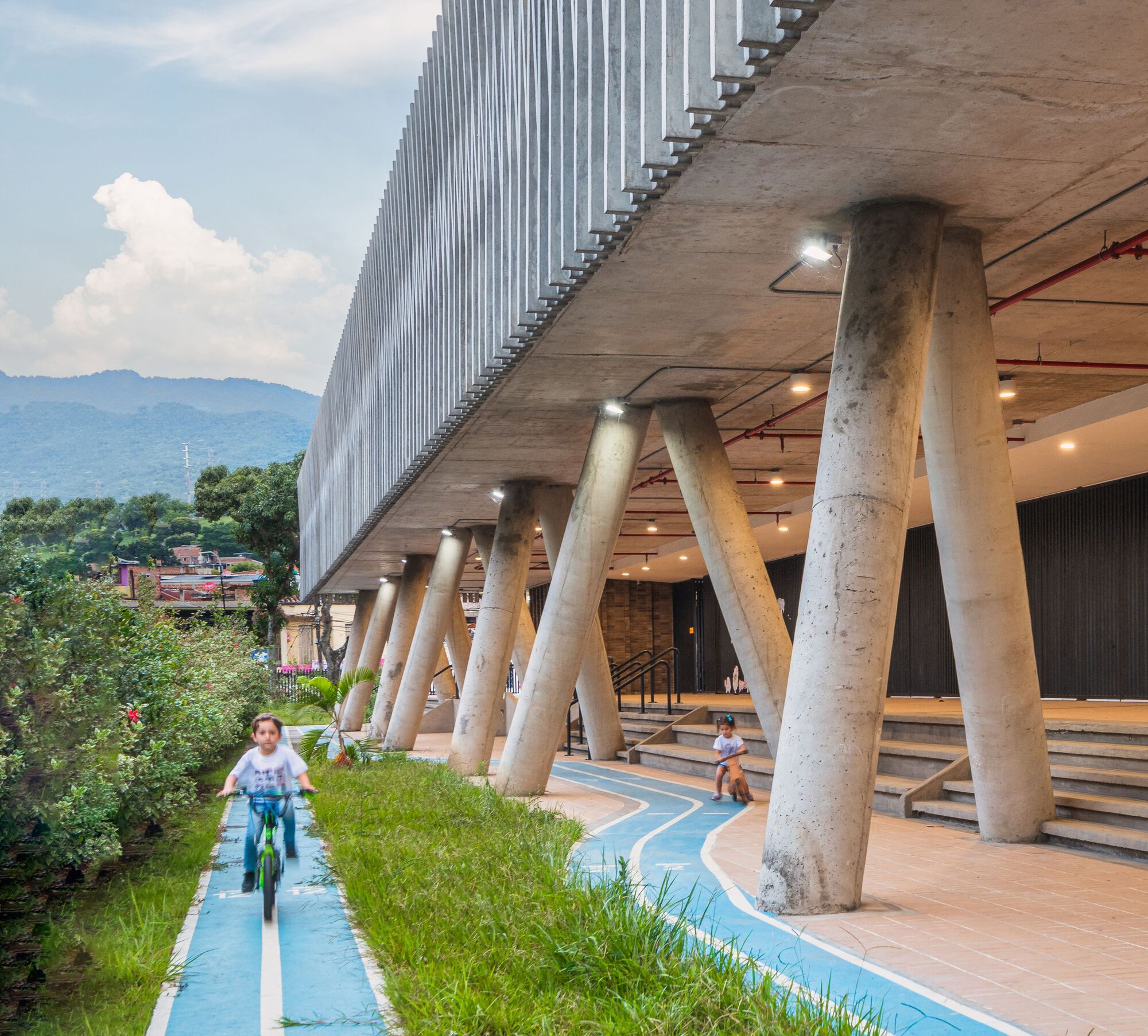 Ground level view of the angled concrete columns, covered walkway, and children on bicycles using the blue path