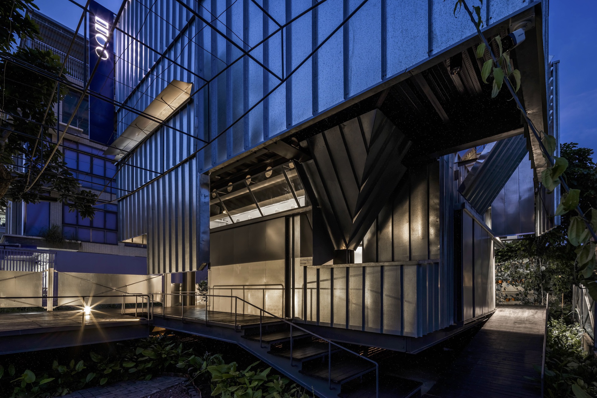 Corner pavilion with folded metal roof and vertical cladding lit from within at twilight