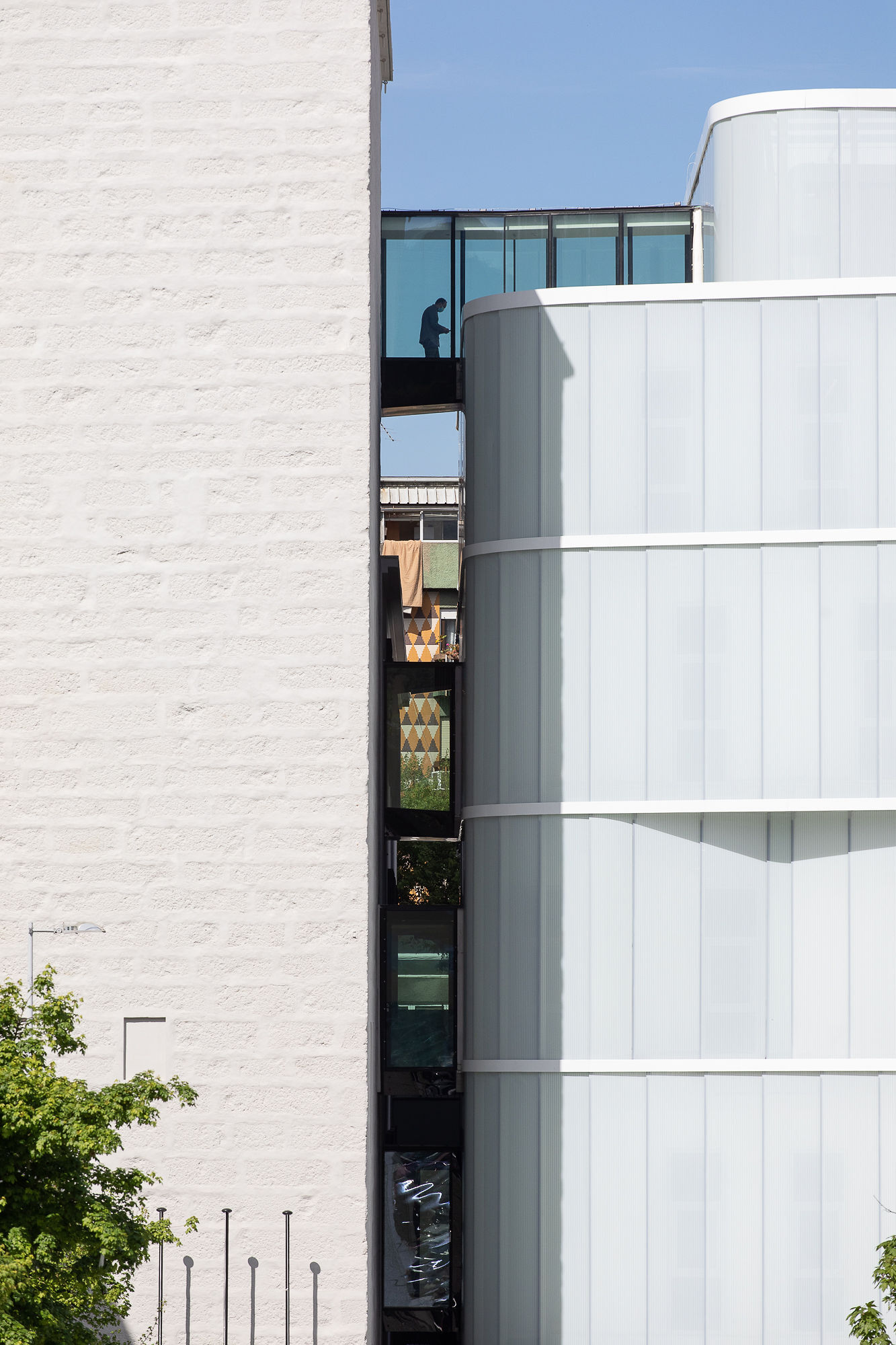 Narrow gap between white brick wall and cylindrical white-paneled facade revealing person on upper balcony