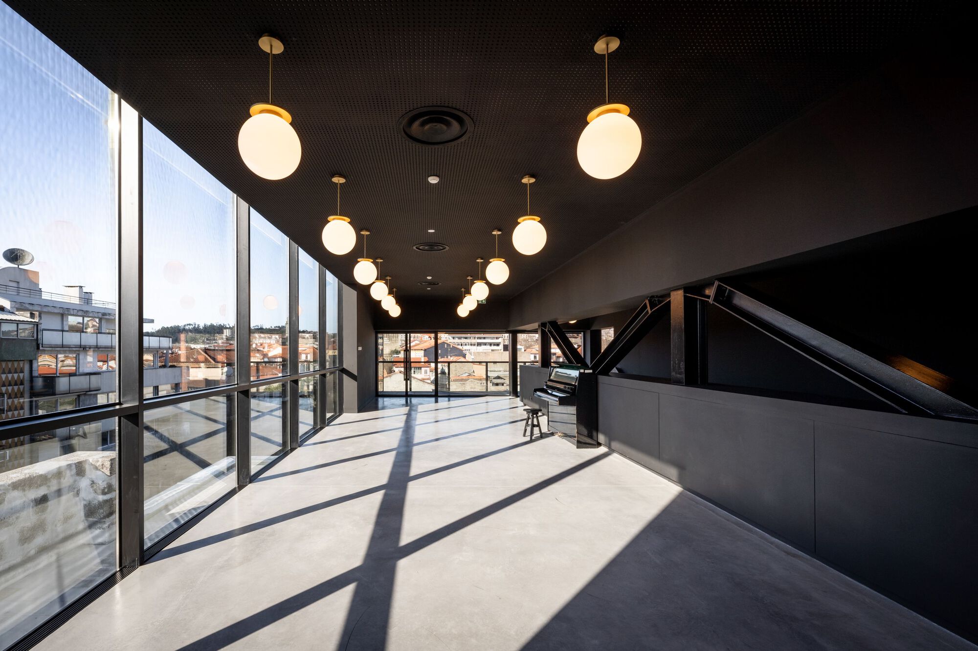 Upper floor corridor with dark perforated ceiling, globe pendant lights, and glazed facade overlooking rooftops