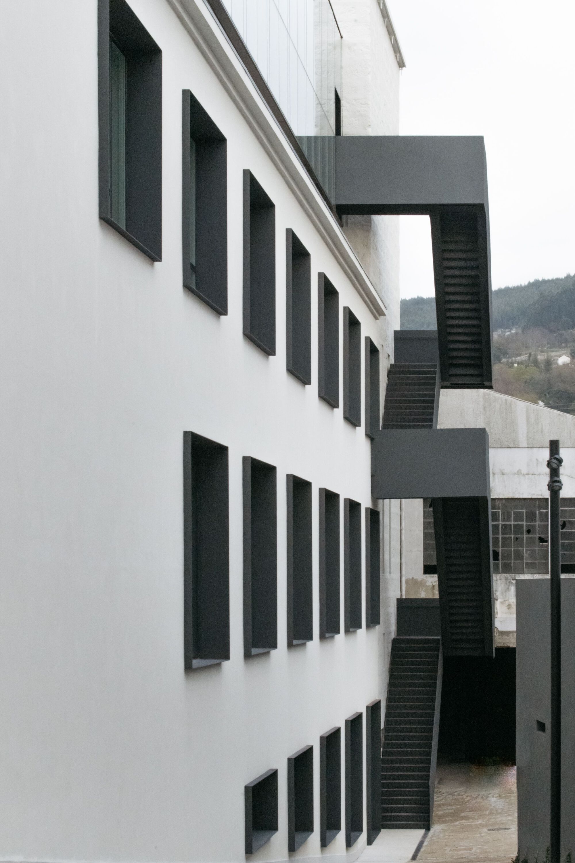 White rendered facade with dark recessed windows and external steel staircase under overcast sky