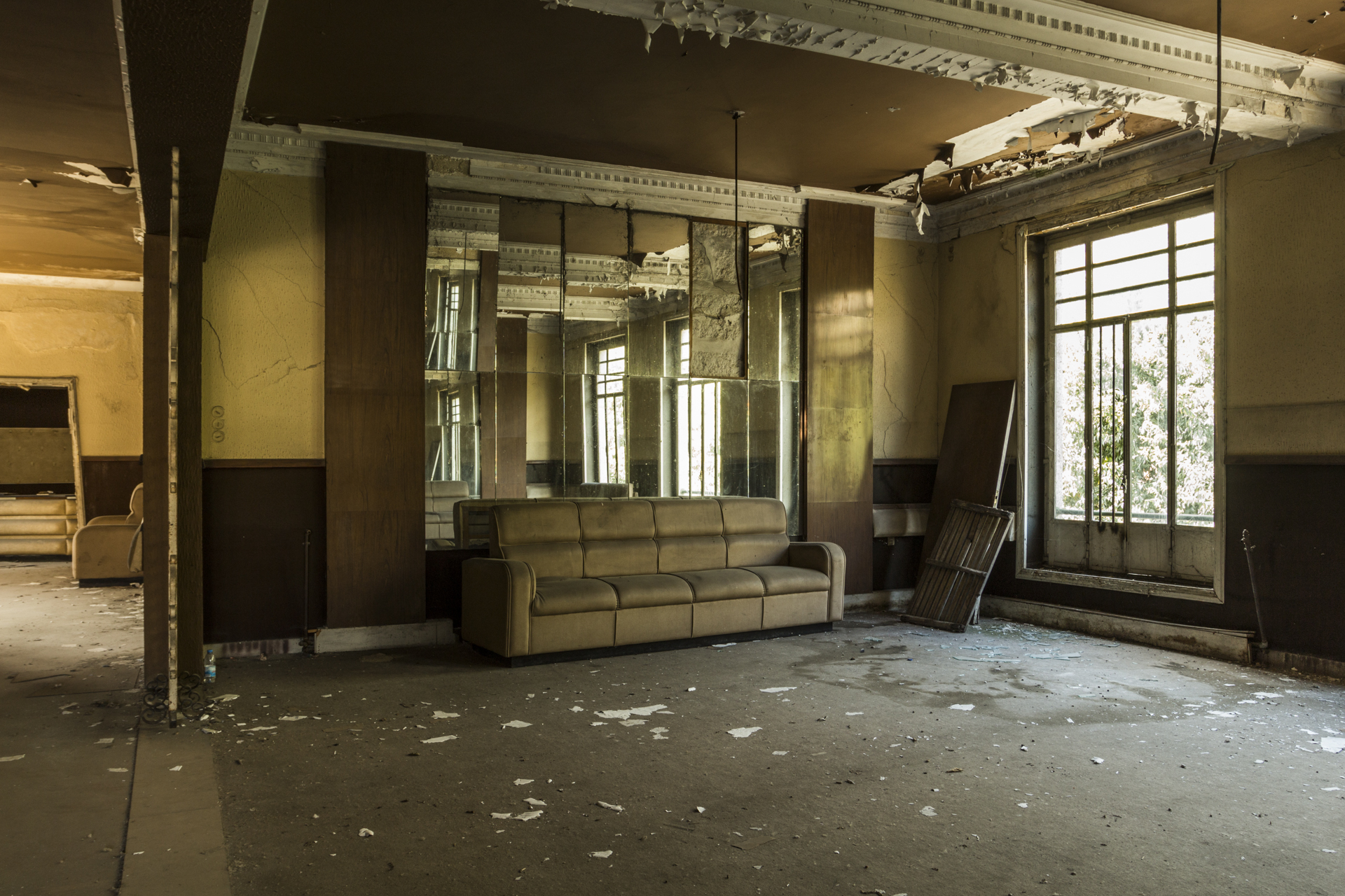 Abandoned interior hall with ornate ceiling moldings, tall windows, and a worn sofa under natural light