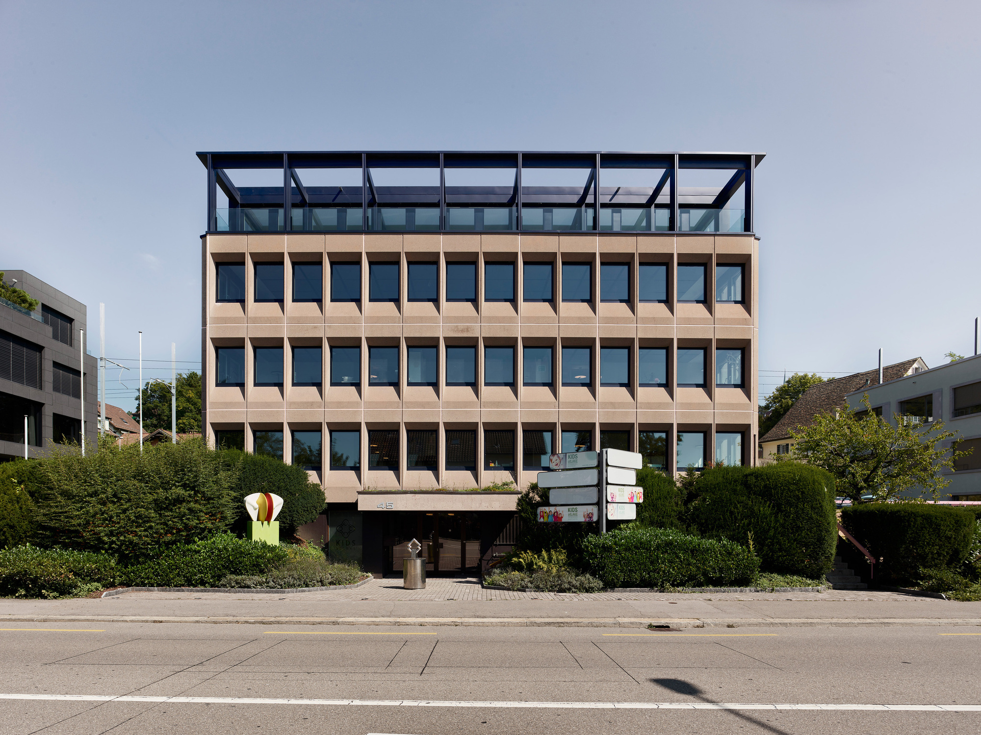 Street view of the layered facade with terracotta panels and a recessed glazed penthouse above green hedges