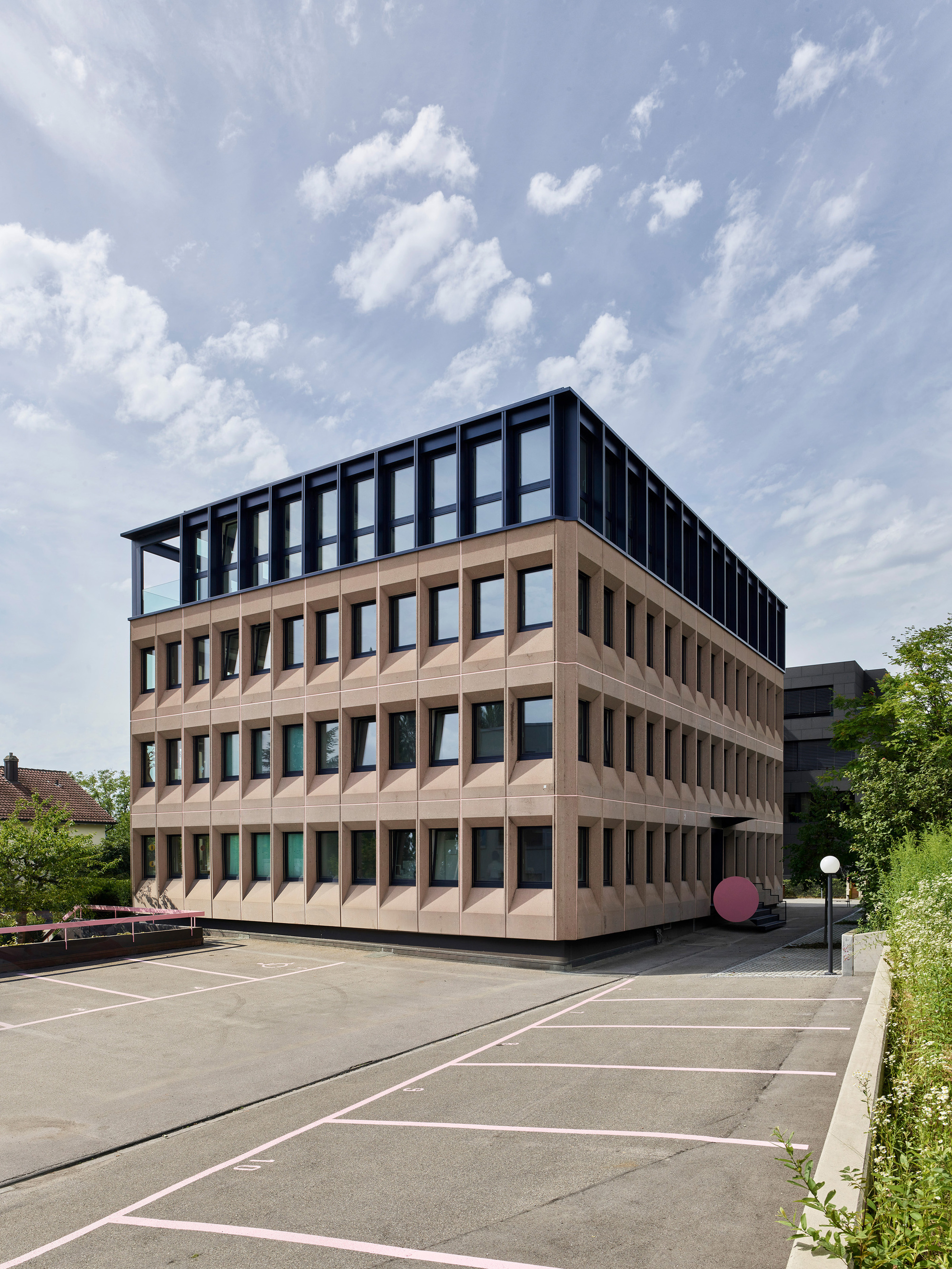 View across the parking area showing the folded terracotta facade and rooftop steel frame under blue skies
