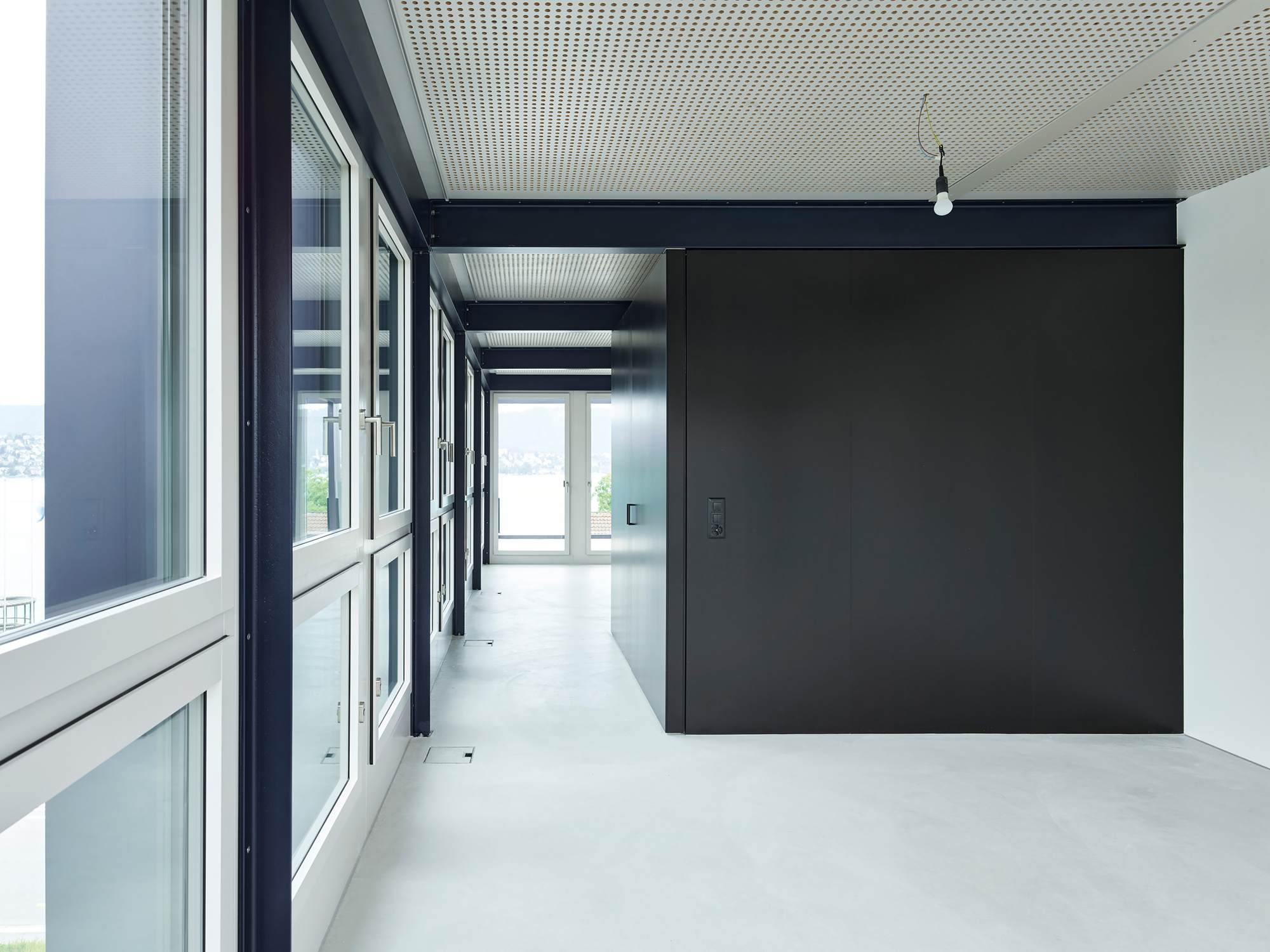 Hallway view along the glazed facade with a dark partition volume and perforated ceiling