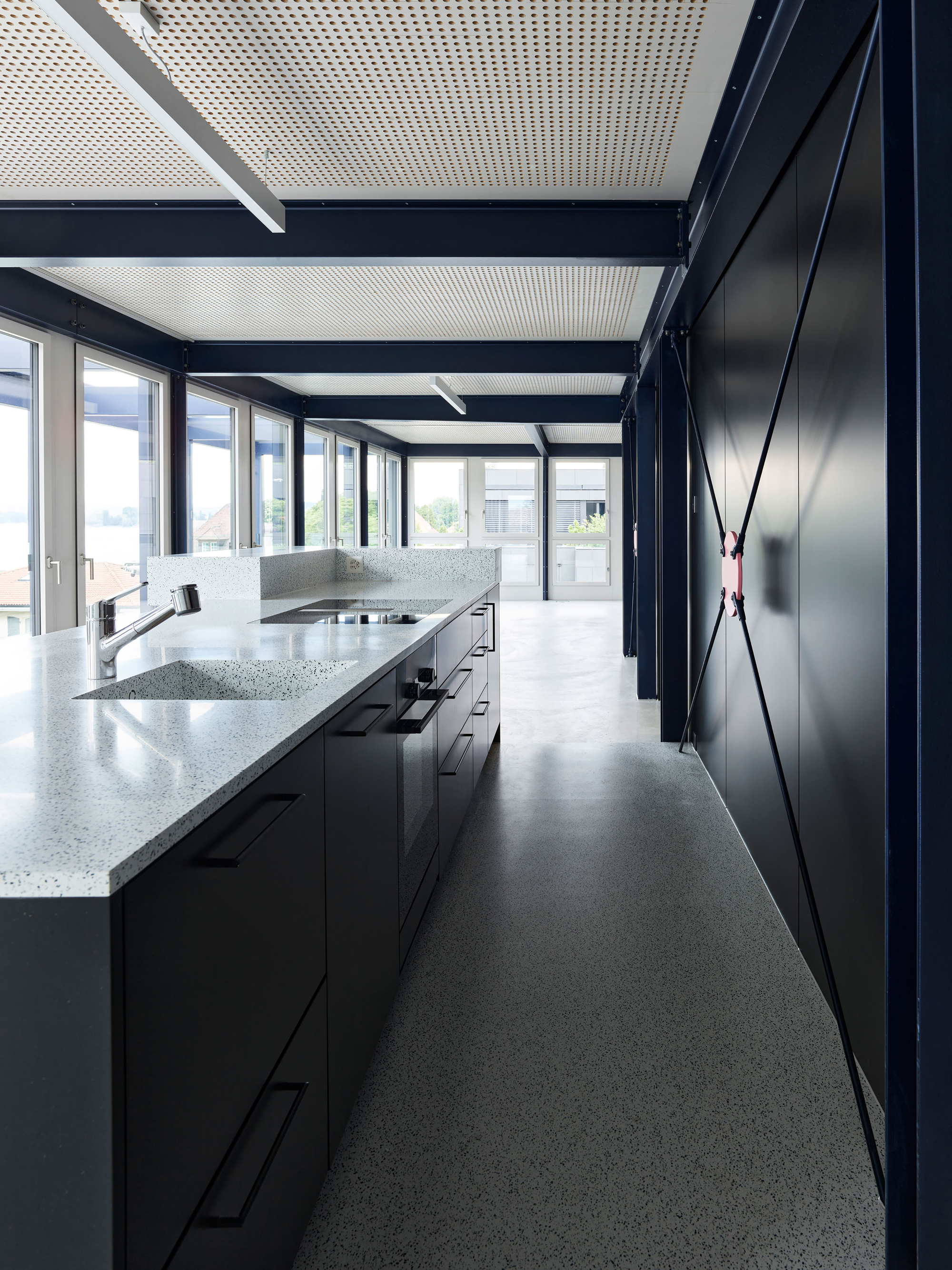 Kitchen with black cabinets, terrazzo countertops, and perforated ceiling panels framed by steel beams