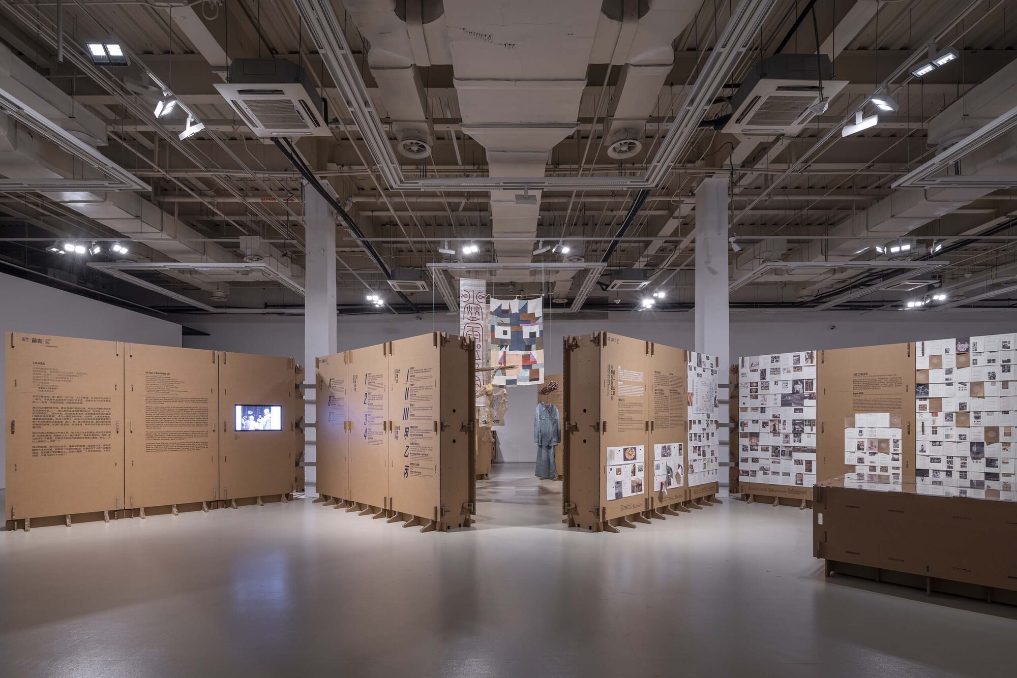 Exhibition space with corrugated cardboard partition walls displaying text and images under exposed ceiling infrastructure