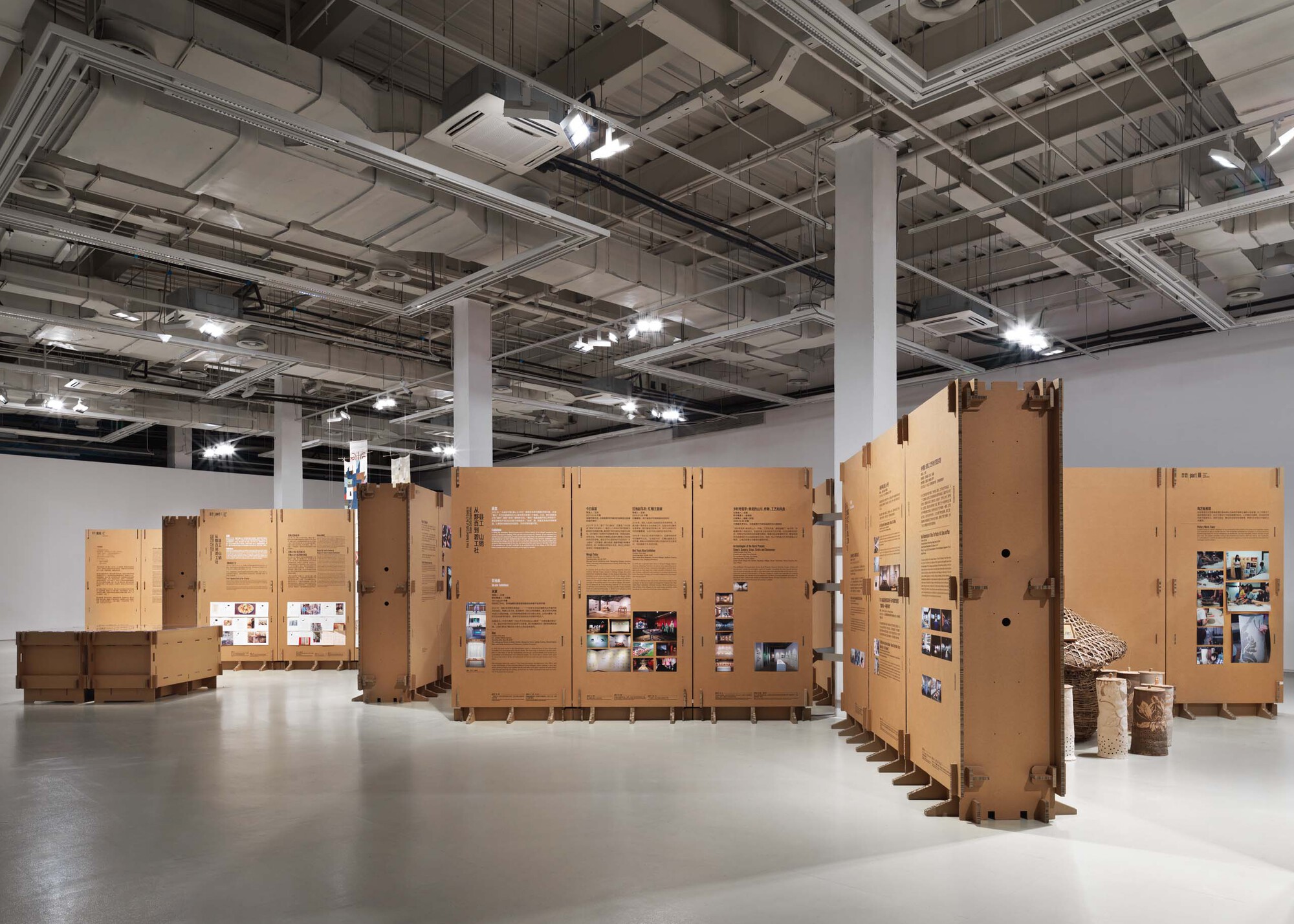 Cardboard exhibition walls arranged in a serpentine configuration with cylindrical woven elements visible at right