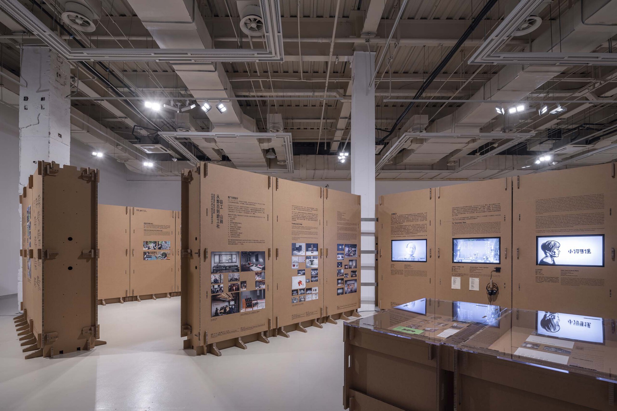View across the installation showing cardboard walls with embedded video screens and photograph collages