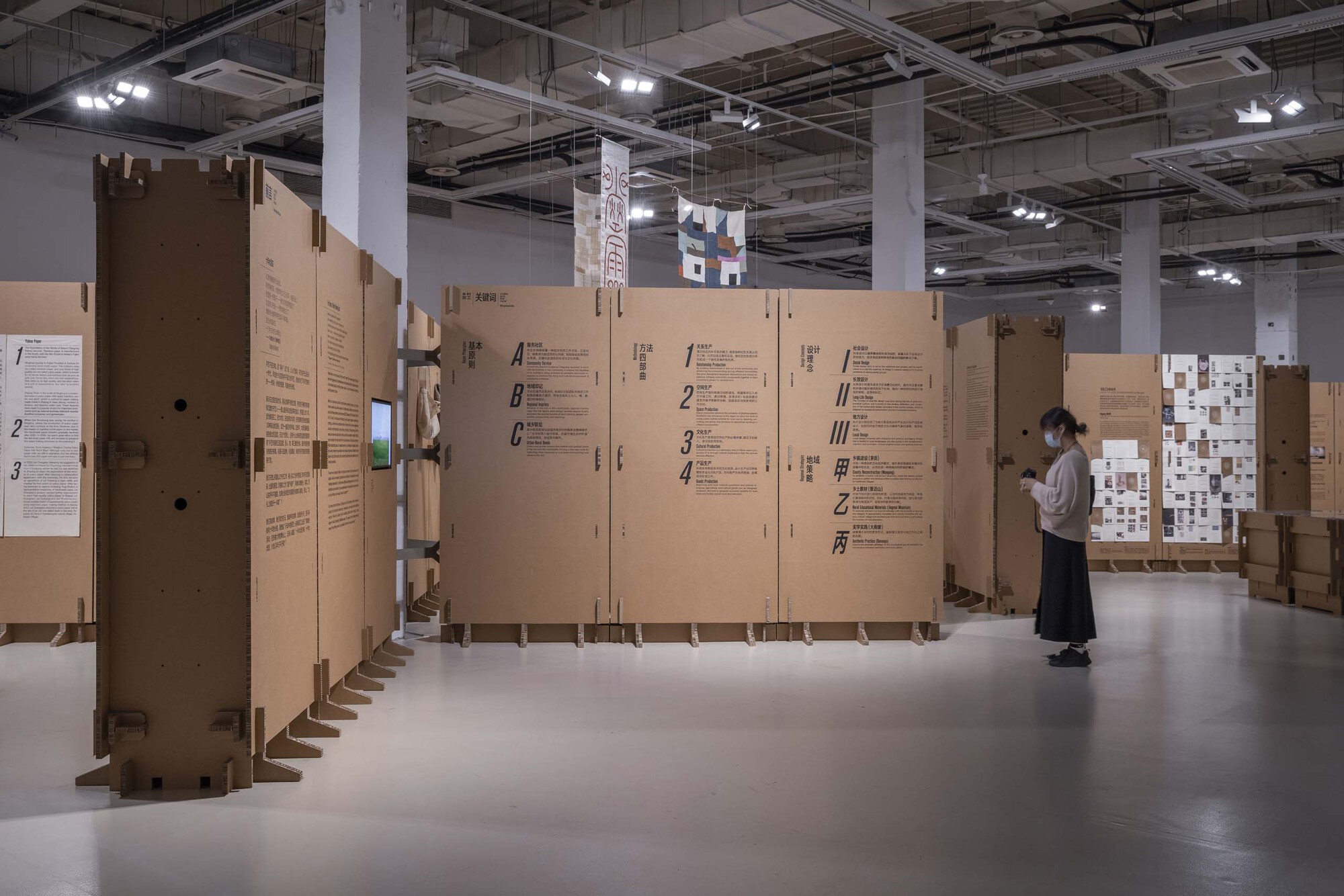 Exhibition space with corrugated cardboard partition walls displaying text panels under exposed ceiling infrastructure