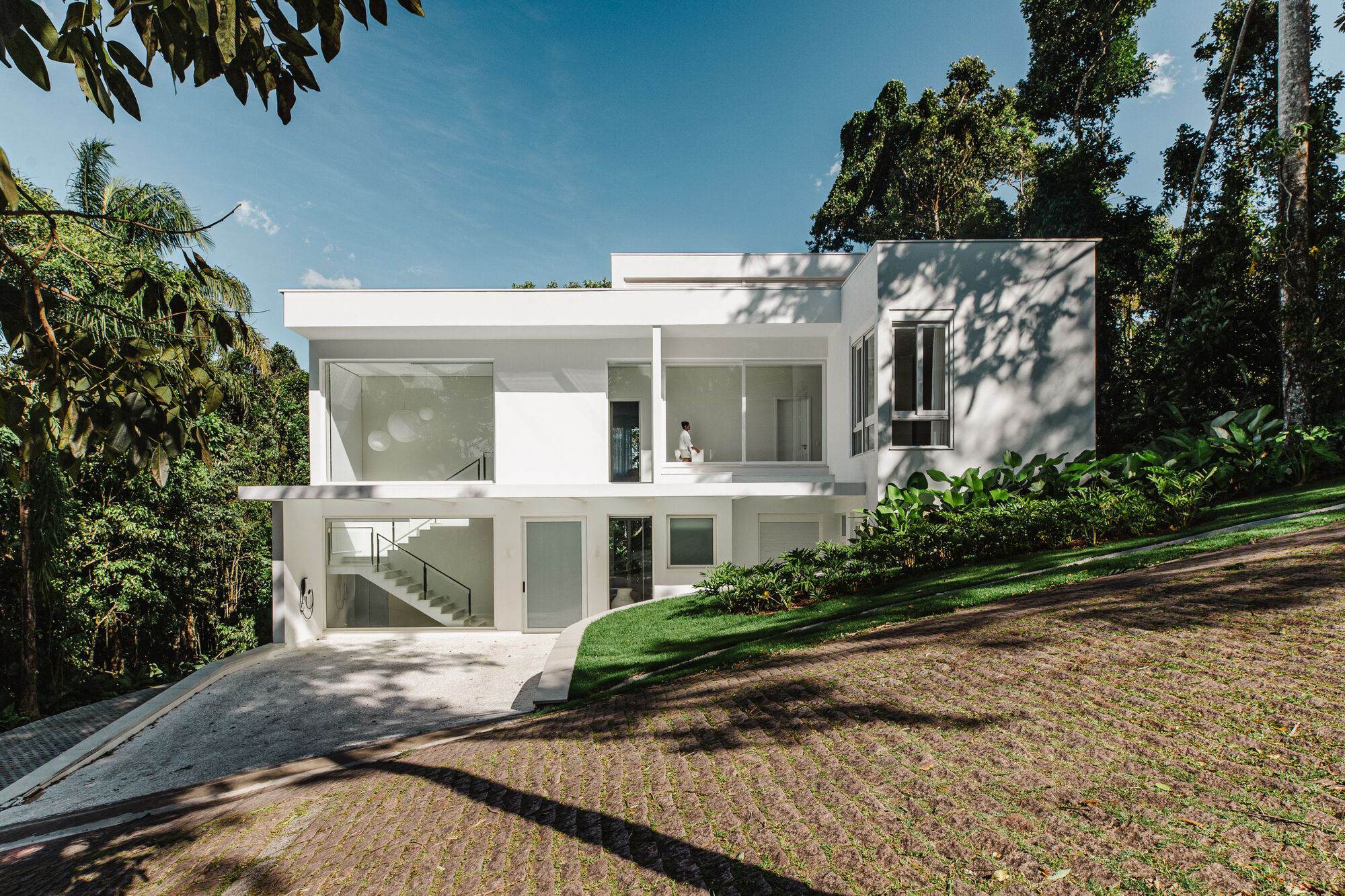 Two-story white volume set into a sloping lawn surrounded by tall trees under afternoon sun