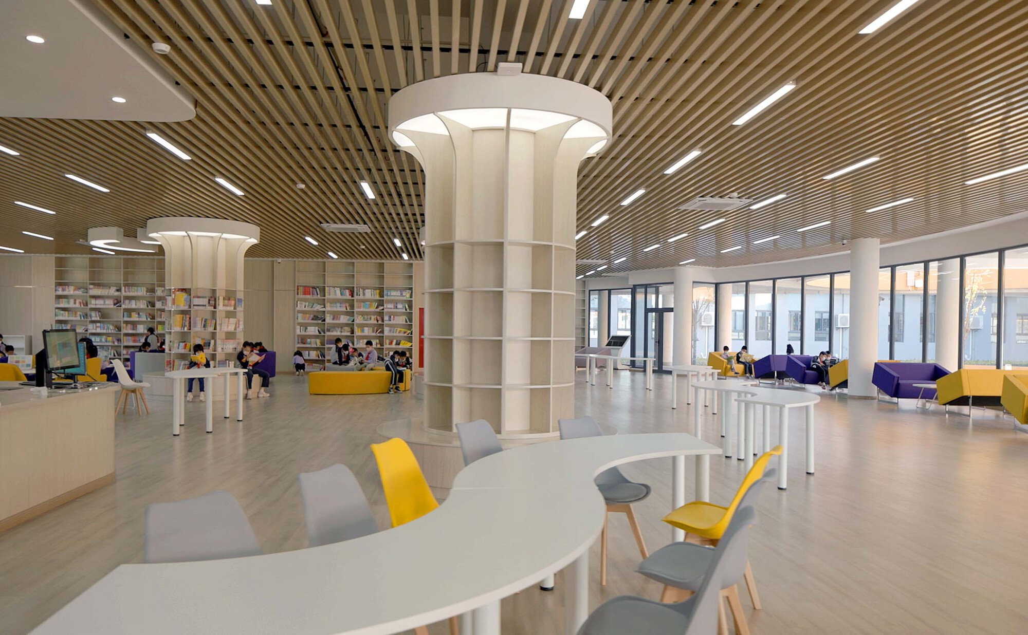 Library interior with slatted timber ceiling, white columns and scattered seating with yellow and grey chairs