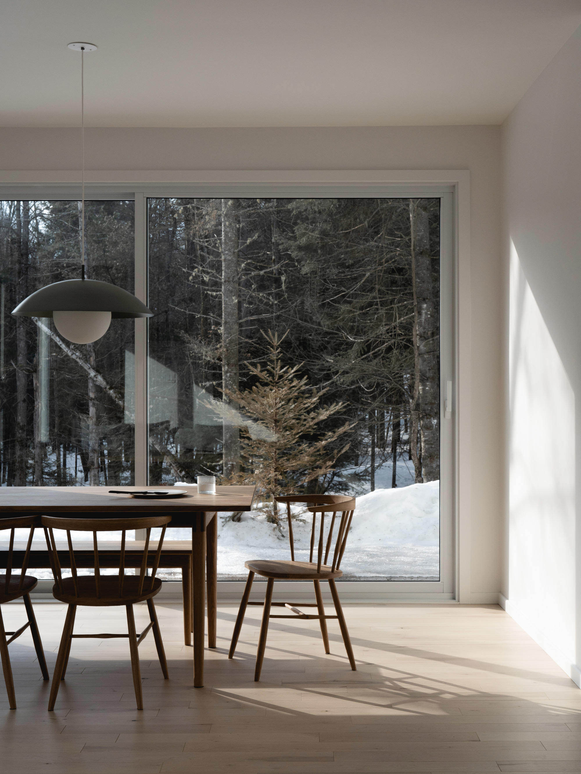 Interior dining area with pendant lamp and timber furniture overlooking the snowy forest through floor-to-ceiling glazing