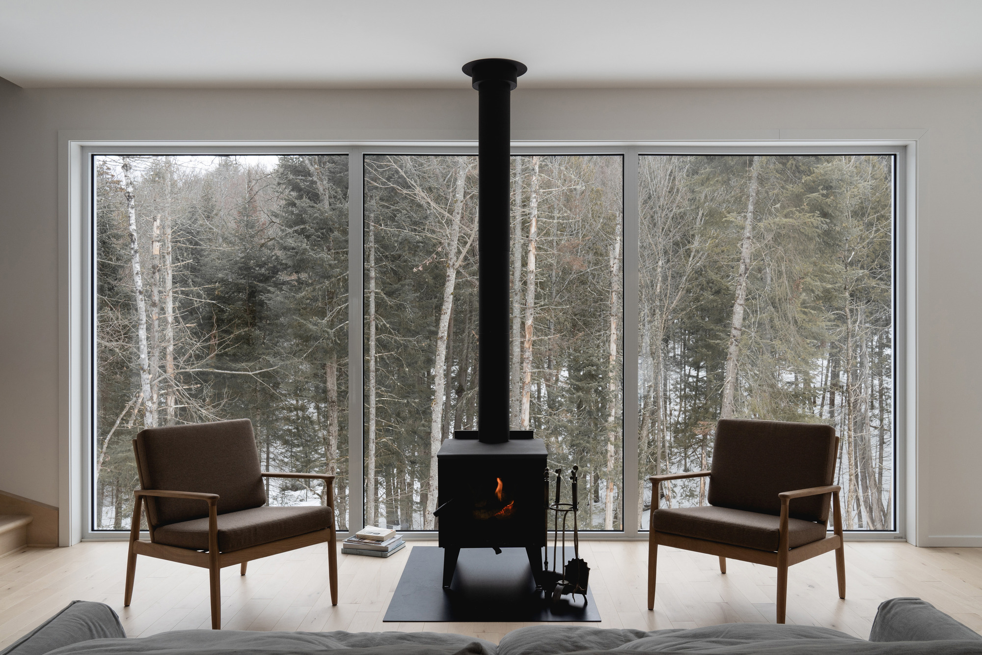 Living room with freestanding black woodstove flanked by two chairs facing the winter forest beyond