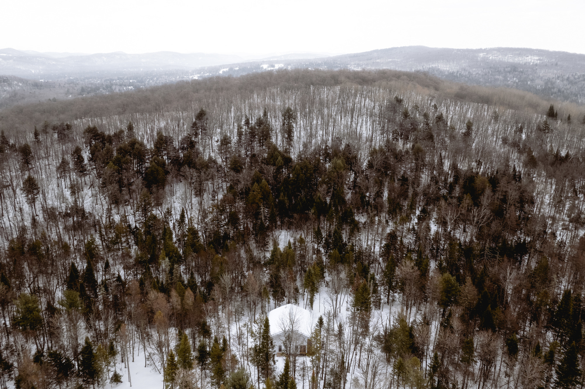 Aerial view of white structure nestled in a snowy mixed forest beneath distant mountain ridges