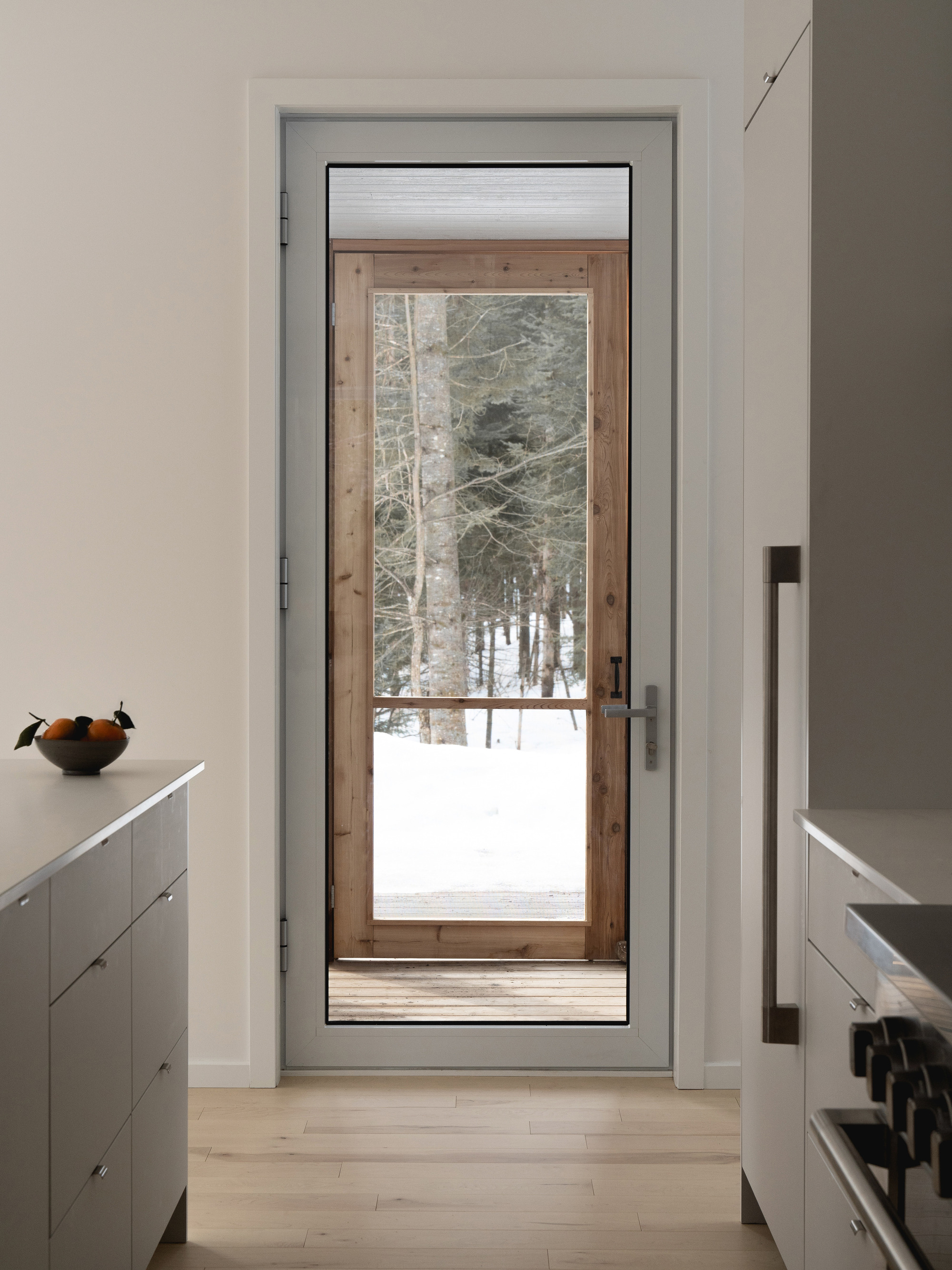 View through a white-framed doorway to a timber-and-glass exterior door with snowy woods beyond