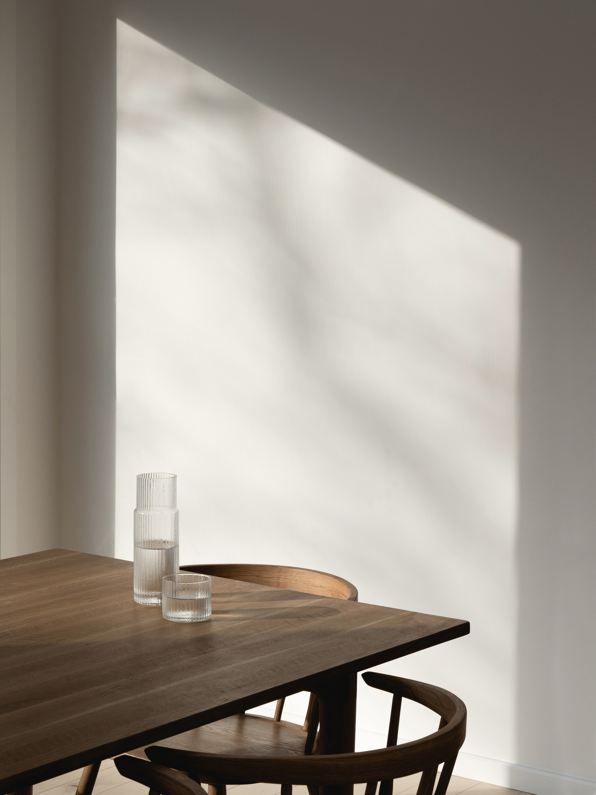 Corner of a walnut dining table with glassware under diagonal shadows cast on white walls