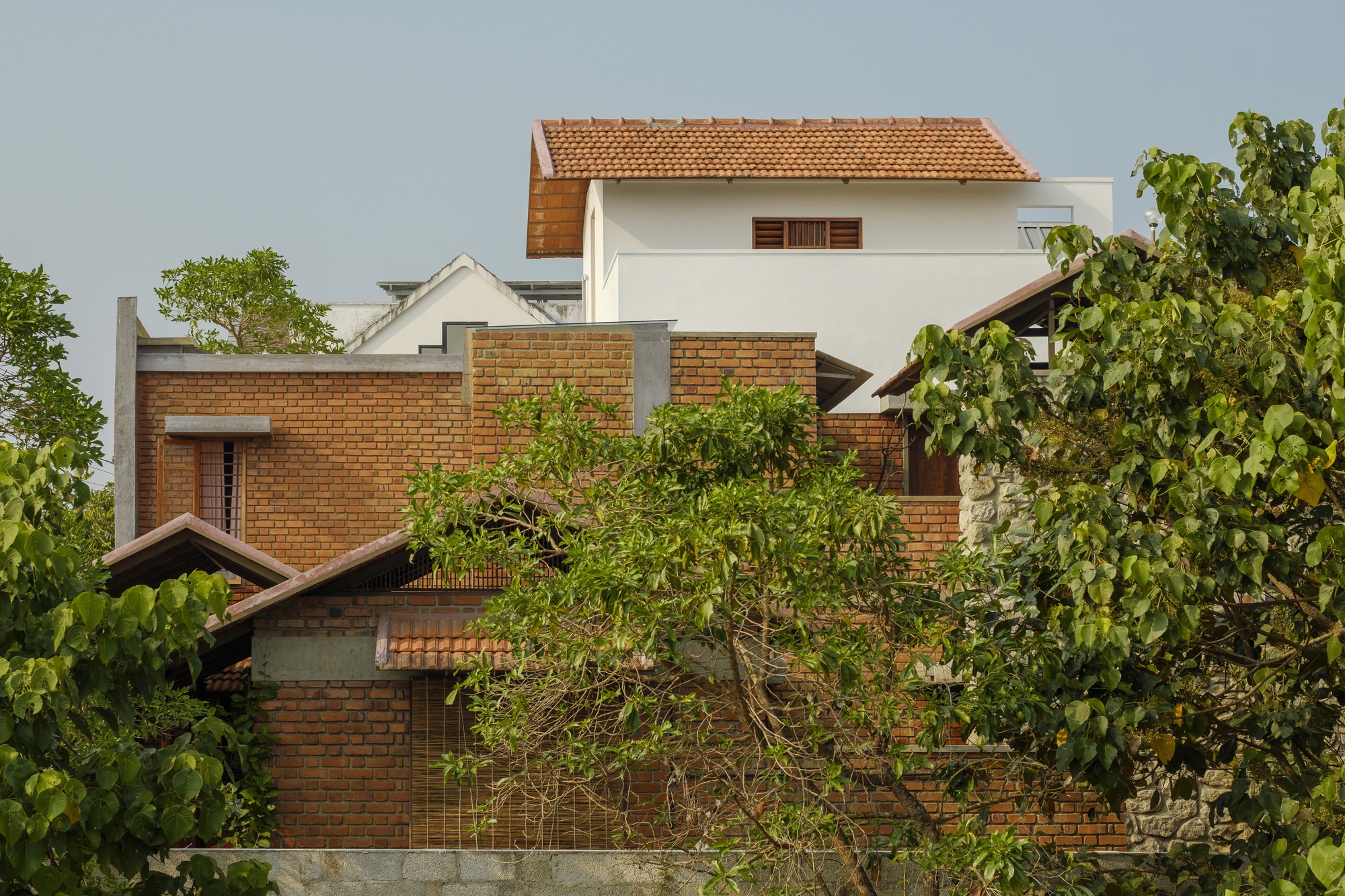 Layered brick volumes with clay tile roofs rising above surrounding trees and foliage