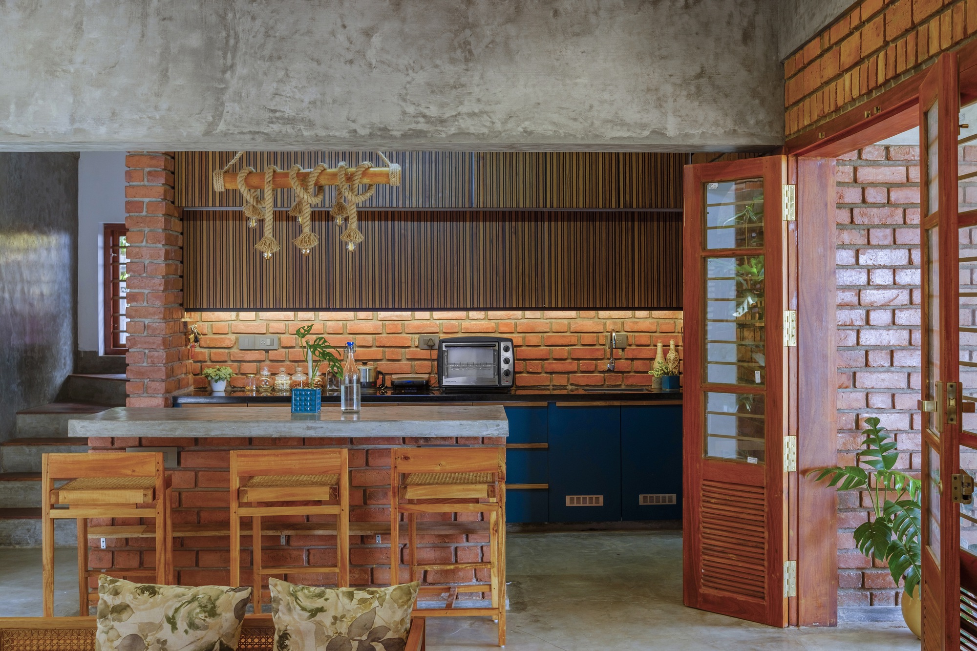 Kitchen with exposed brick island, timber slatted upper cabinets and brass pendant lights beneath a raw concrete ceiling