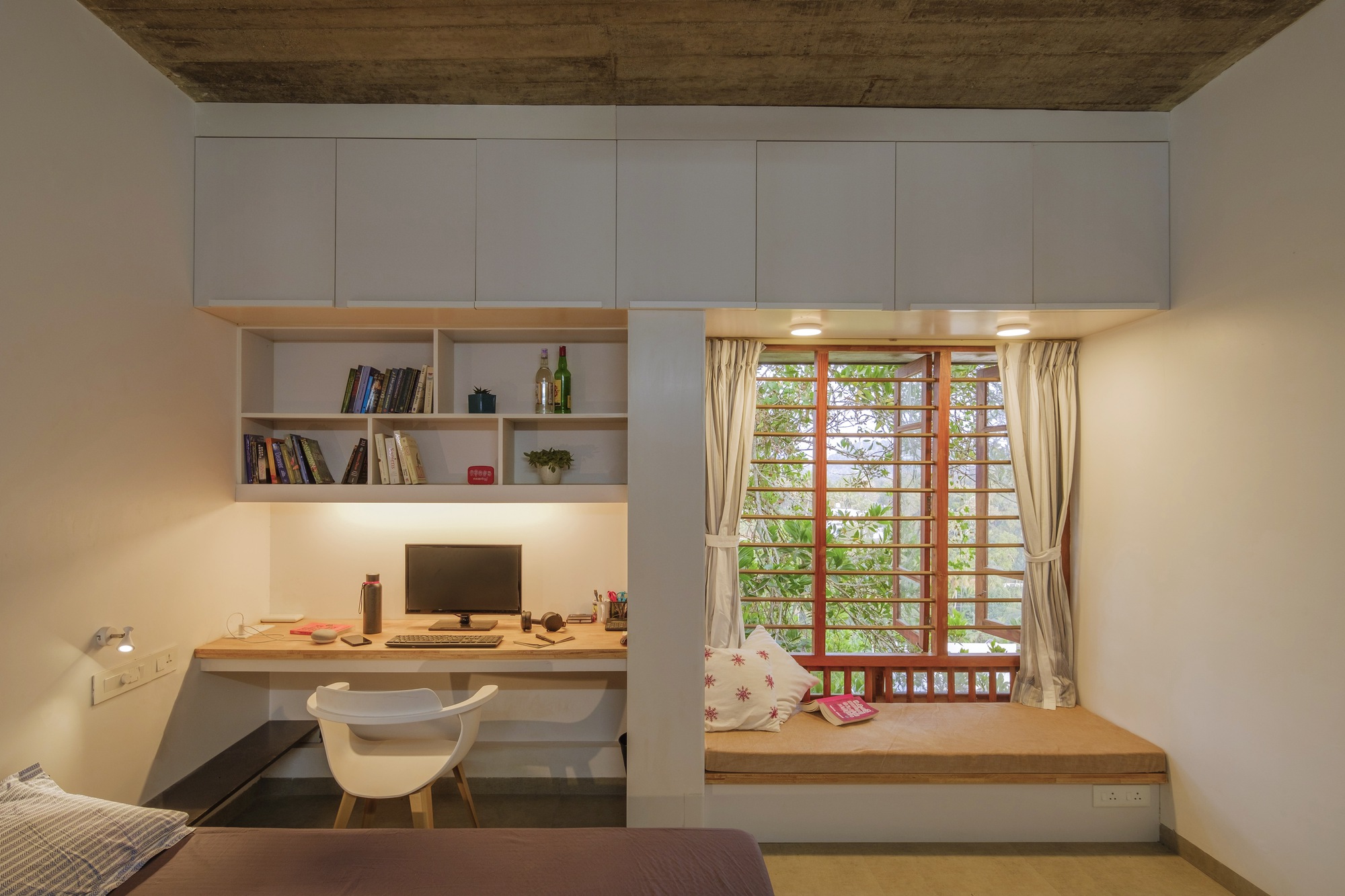 Bedroom workspace with built-in desk, white storage wall and window seat overlooking green foliage through latticed timber shutters