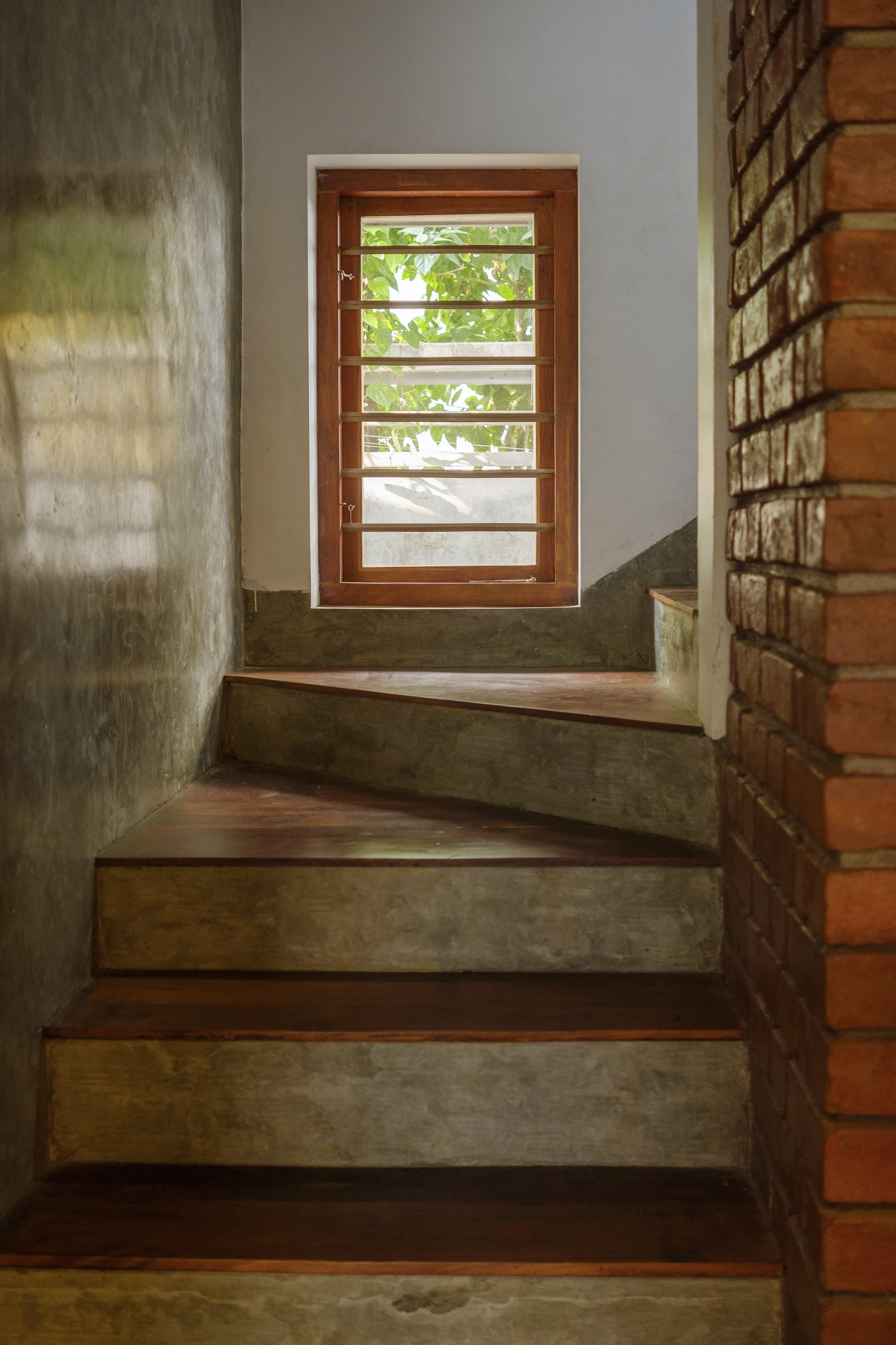 Interior staircase with alternating wood and concrete treads leading to a horizontal louver door with green foliage visible beyond
