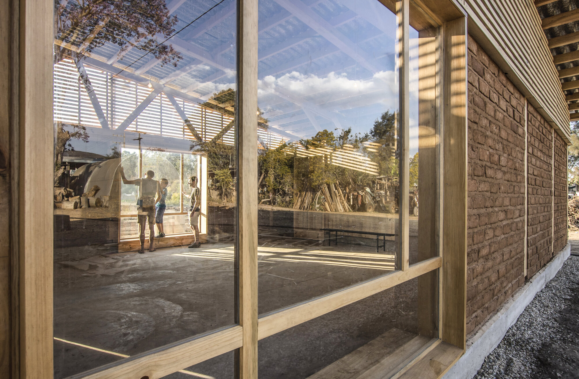Glass-walled corridor with timber frame and slatted roof overhang casting striped shadows on concrete floor