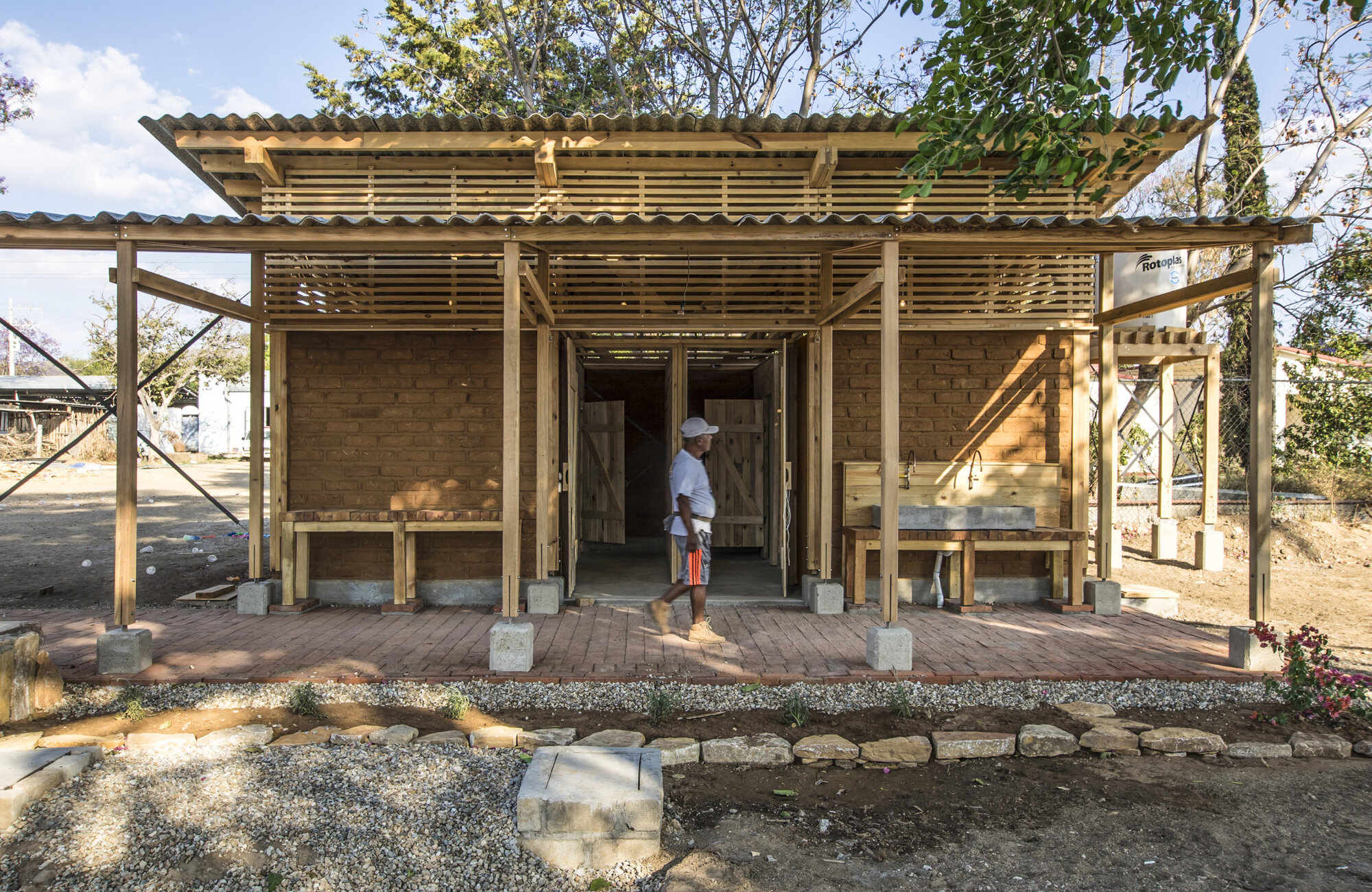 Front facade of a timber-frame pavilion with corrugated metal roof and a person standing in the doorway