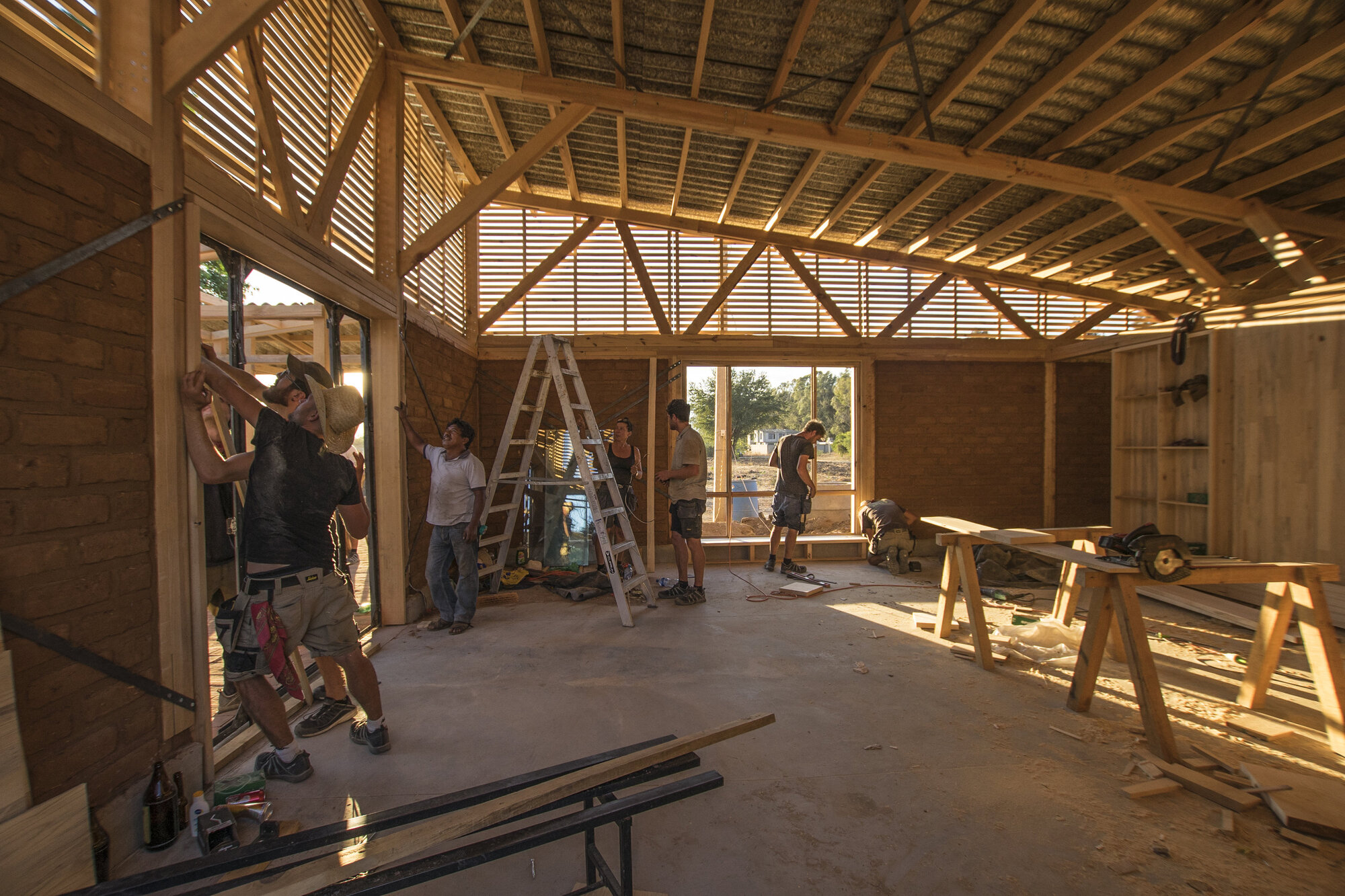 Interior space under construction with exposed timber trusses and workers installing wall panels in afternoon light