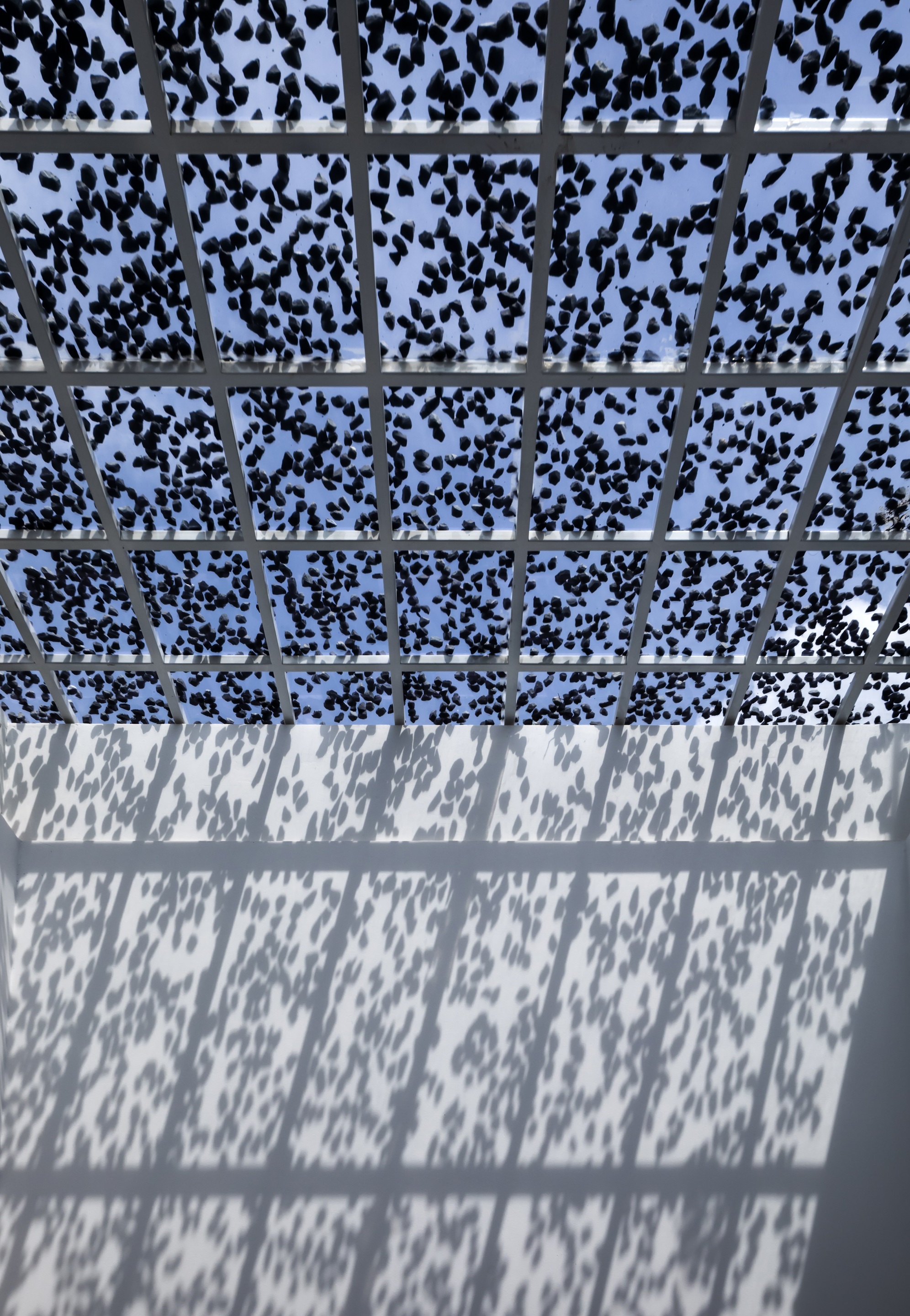 Perforated metal canopy overhead casting dappled shadows on the white wall below in bright sunlight