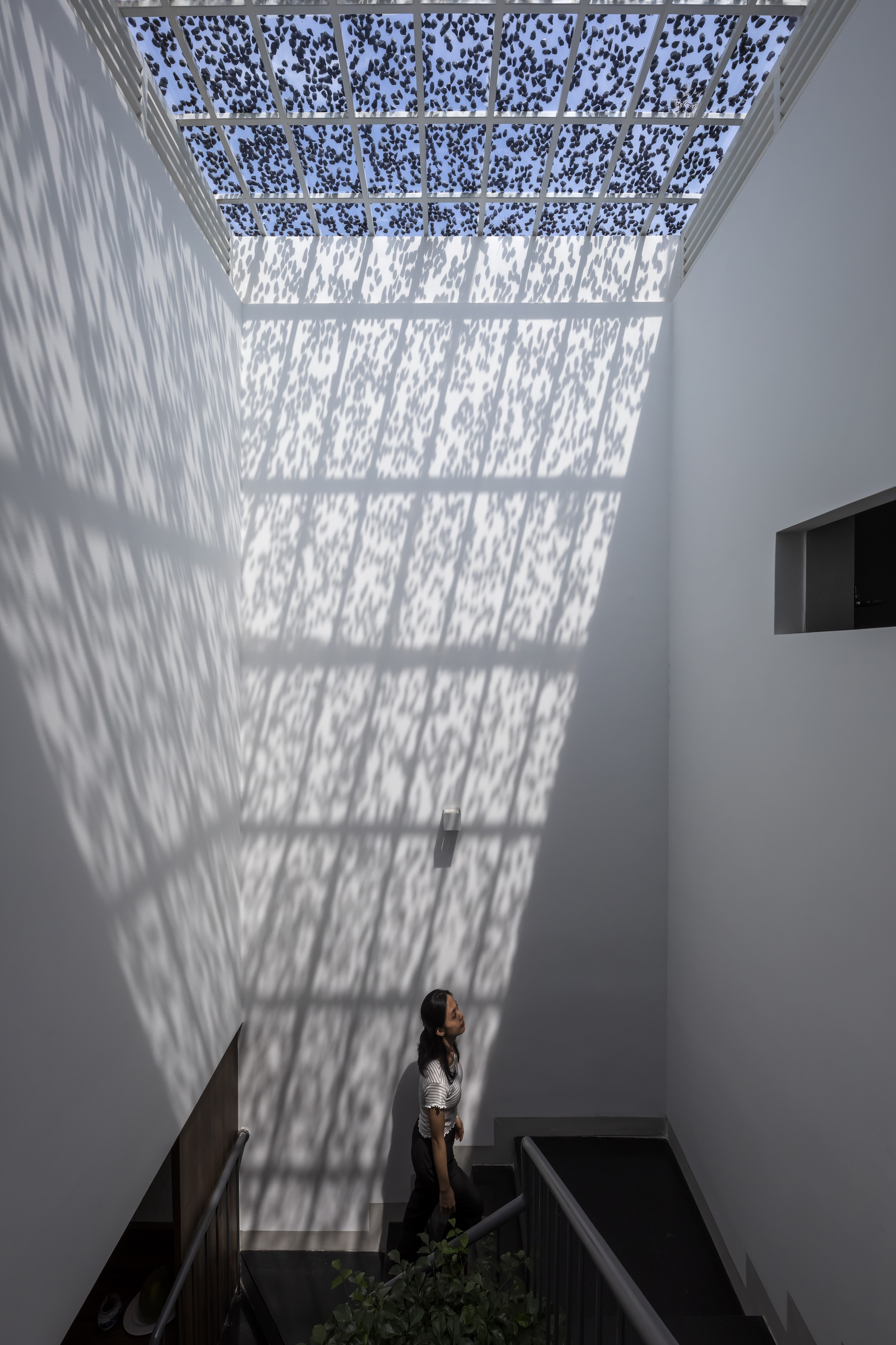Double-height white stairwell with perforated skylight casting dappled shadows on the wall where a woman stands