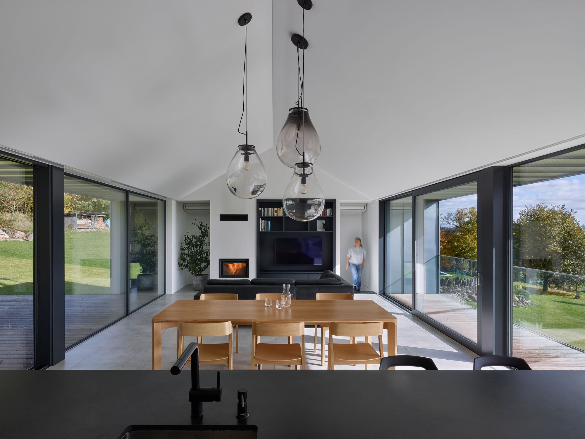 Interior view through dining and living spaces with glass pendant lights and a person visible on the exterior deck