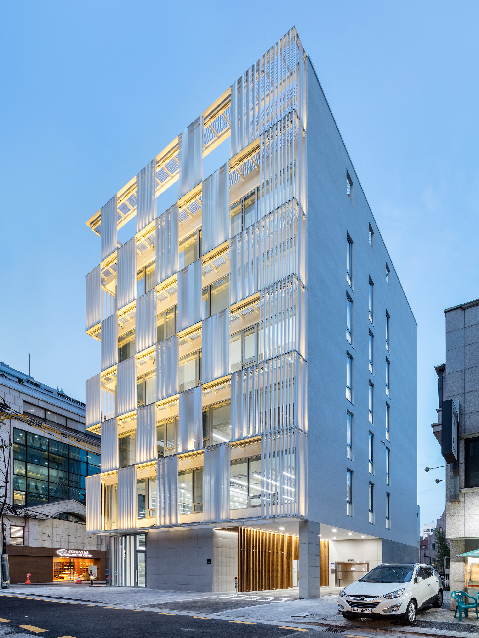 Street view of the white louvered tower facade illuminated at dusk with parked cars in front