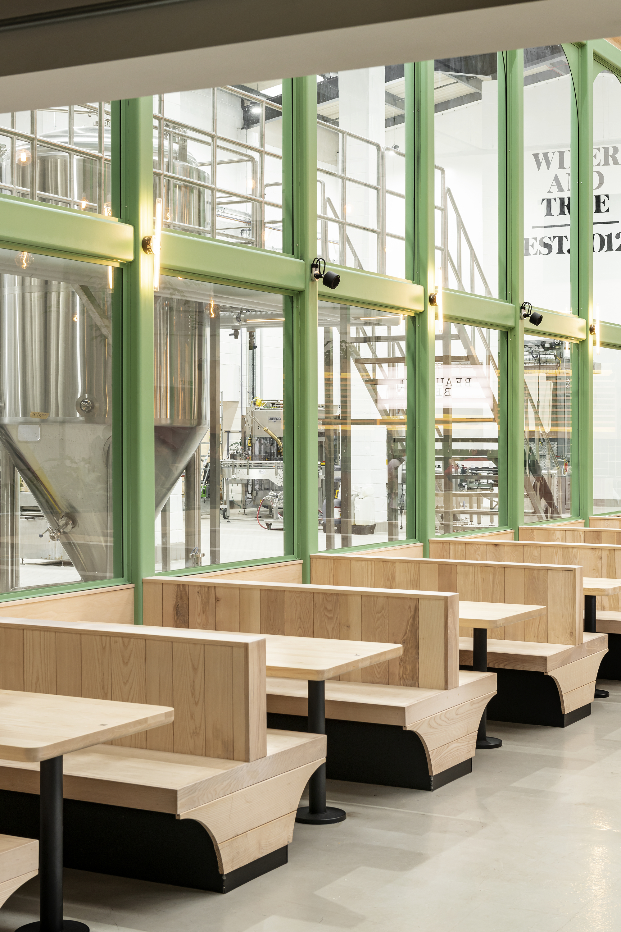 Row of pale timber booth seats beside green-framed glazing with views into the brewery production area