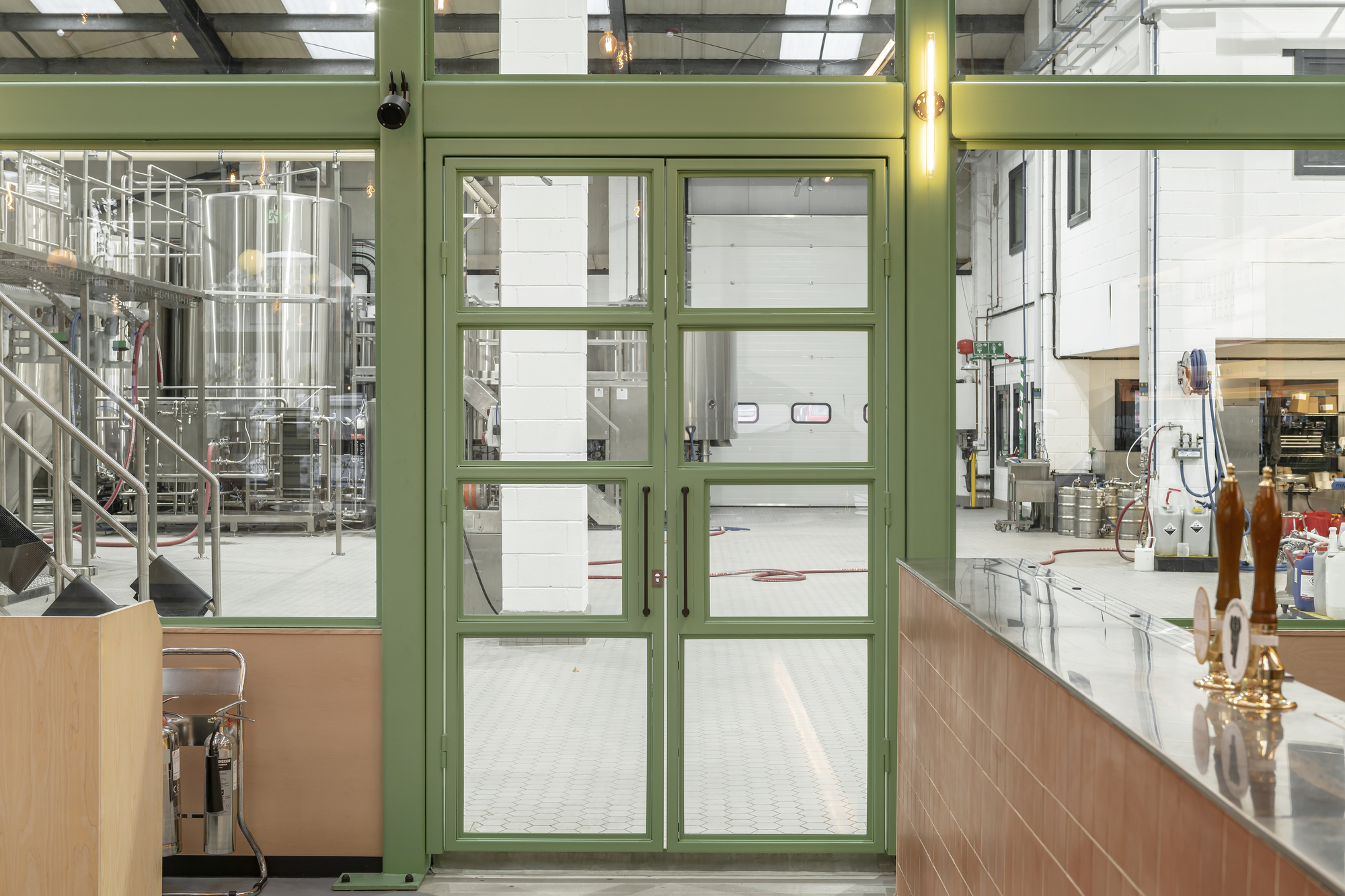 Green framed glass partition separating dining area from brewery equipment and stainless steel tanks beyond