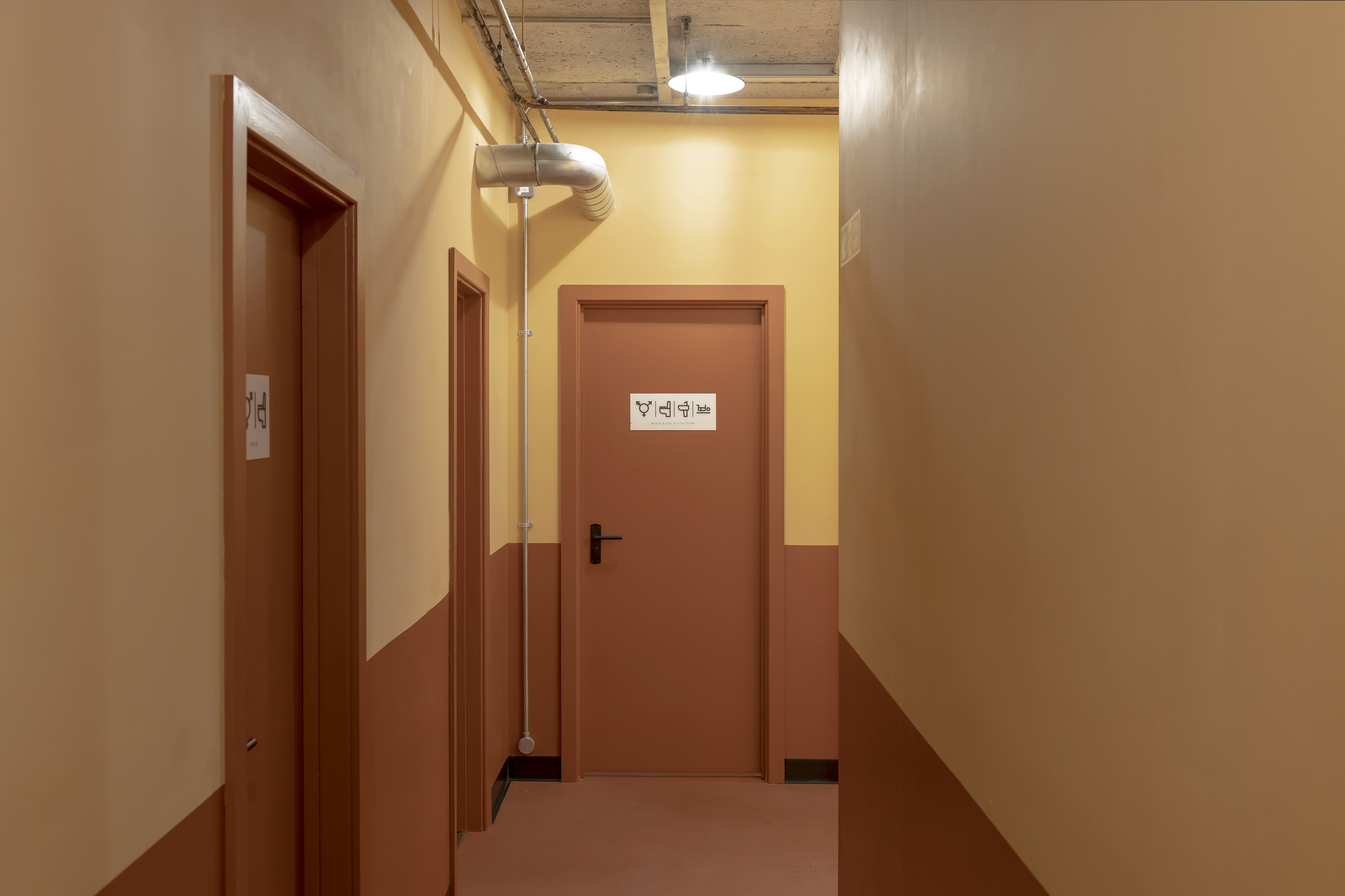 Corridor with terracotta and cream painted walls leading to a restroom door beneath exposed services