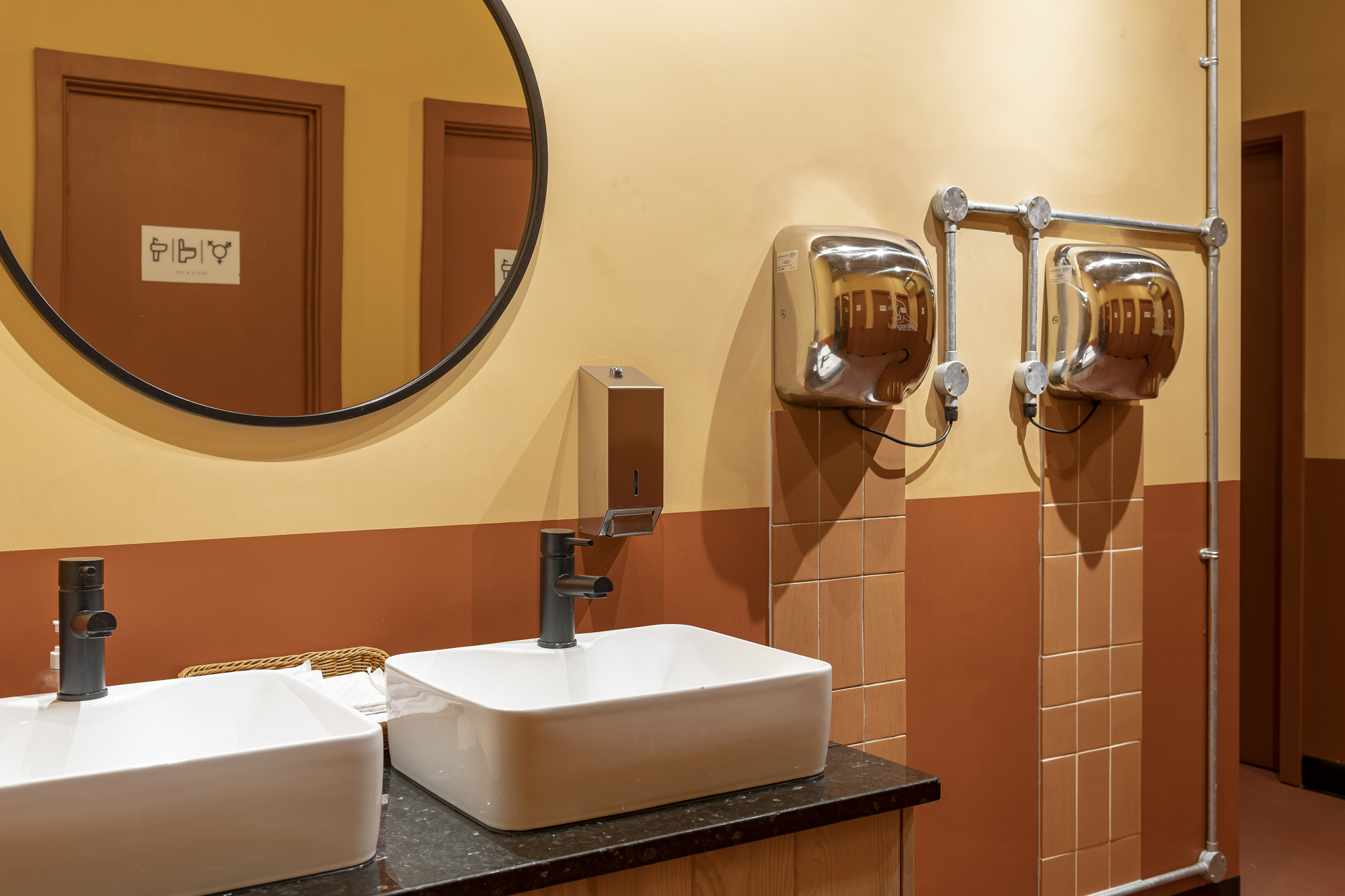 Restroom vanity with twin white basins and chrome hand dryers against terracotta tiled walls