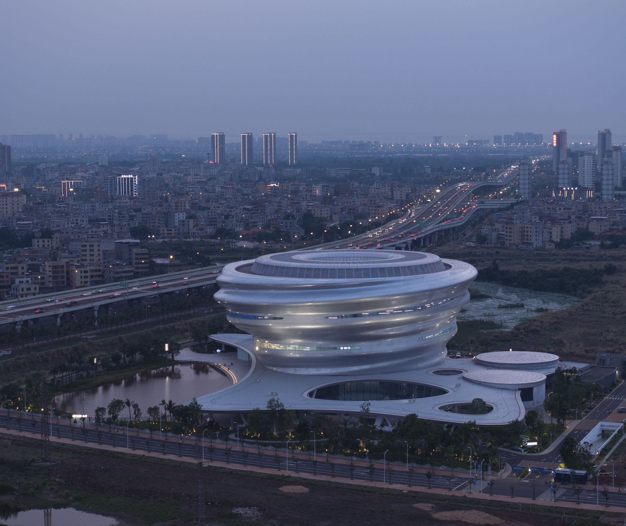 Aerial view of the spiraling ribbed volume beside a highway at dusk with city lights illuminating the background