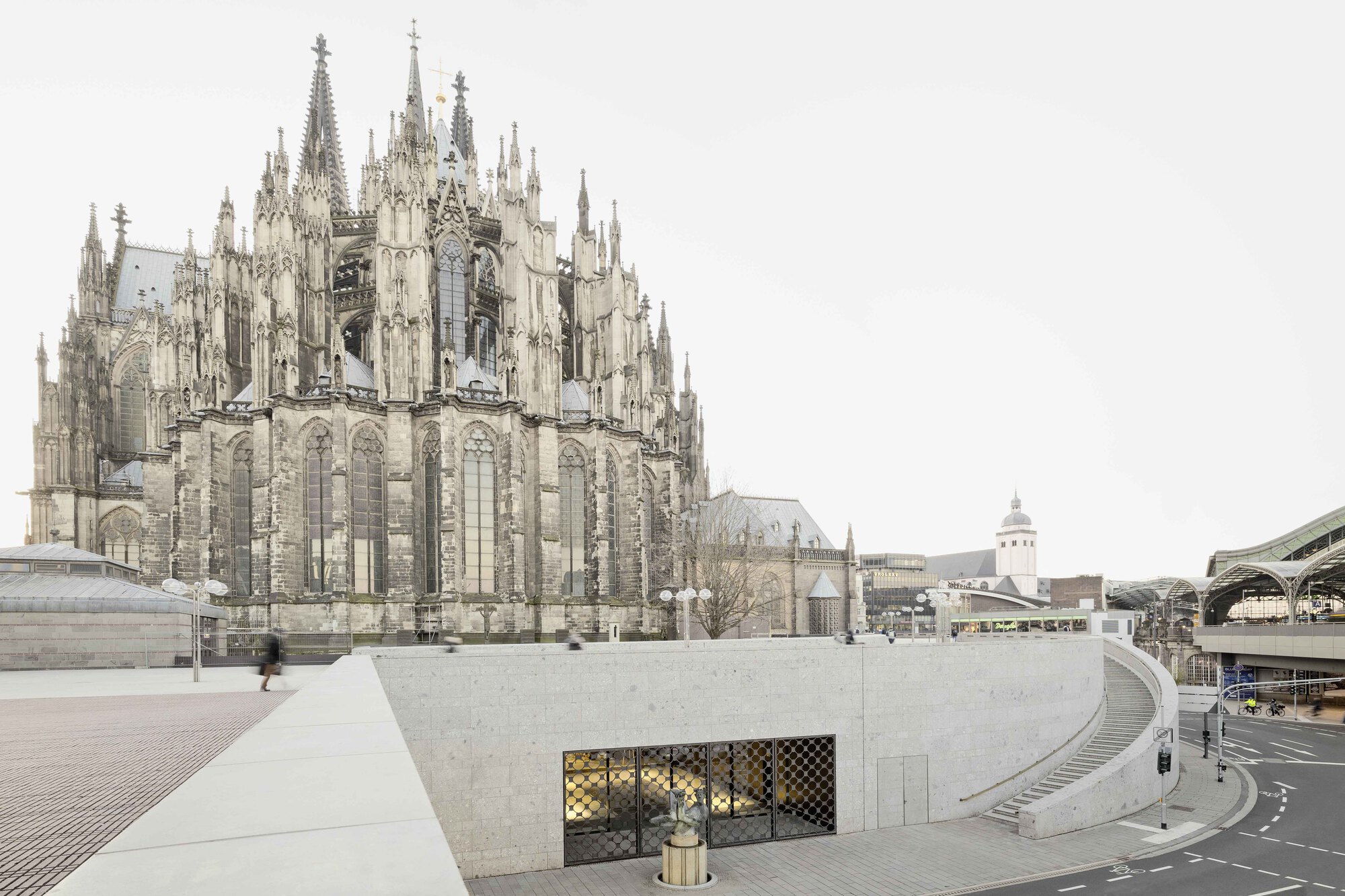 Curved concrete plaza with mesh-screened opening set into the retaining wall below the cathedral