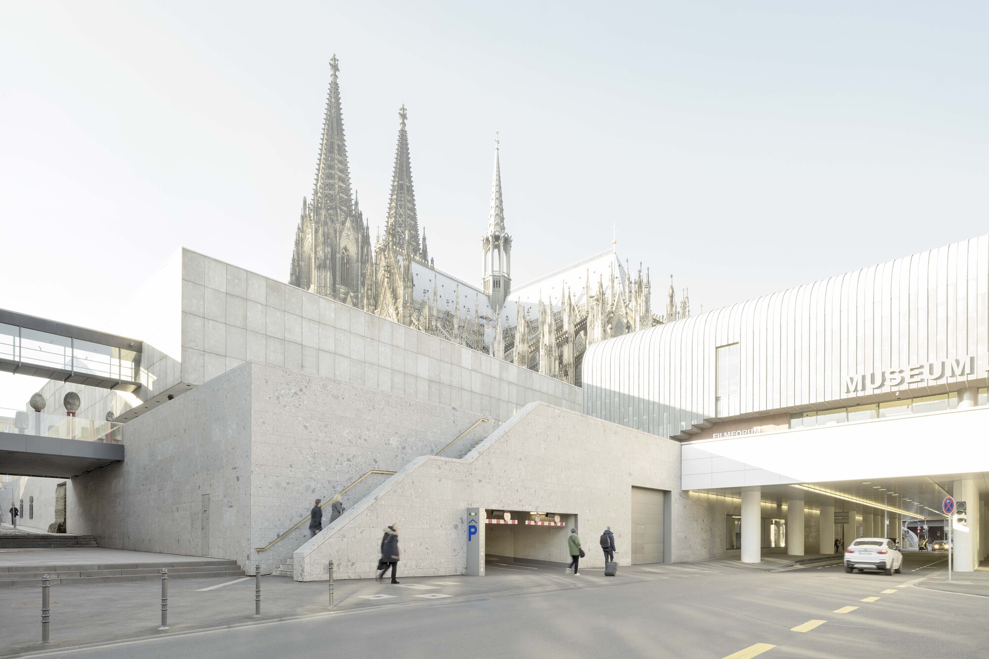 Angled concrete volumes creating a sunken entry with exterior stairs and scattered pedestrians