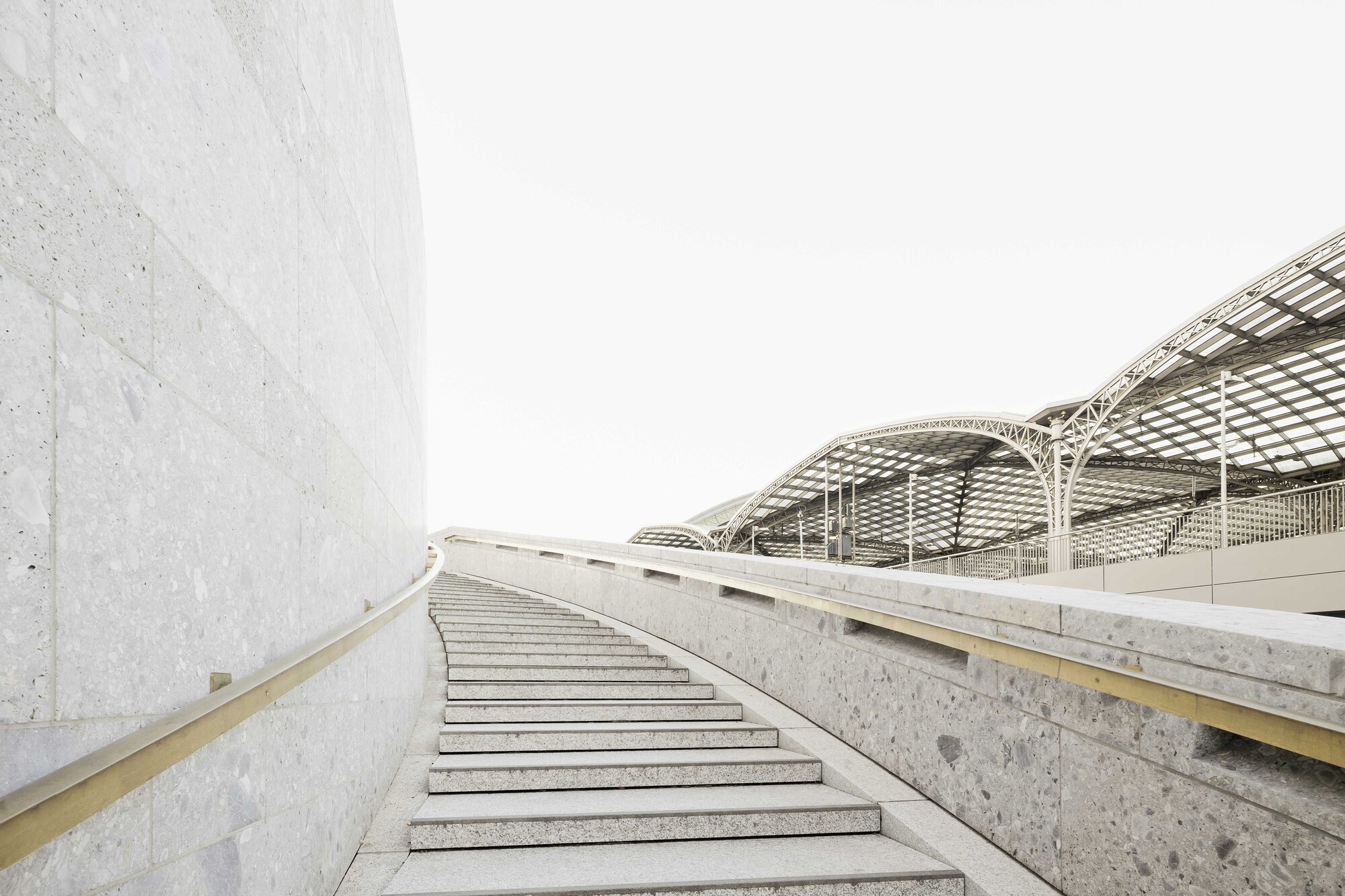 Concrete staircase curving upward alongside a cast wall under a lattice roof canopy