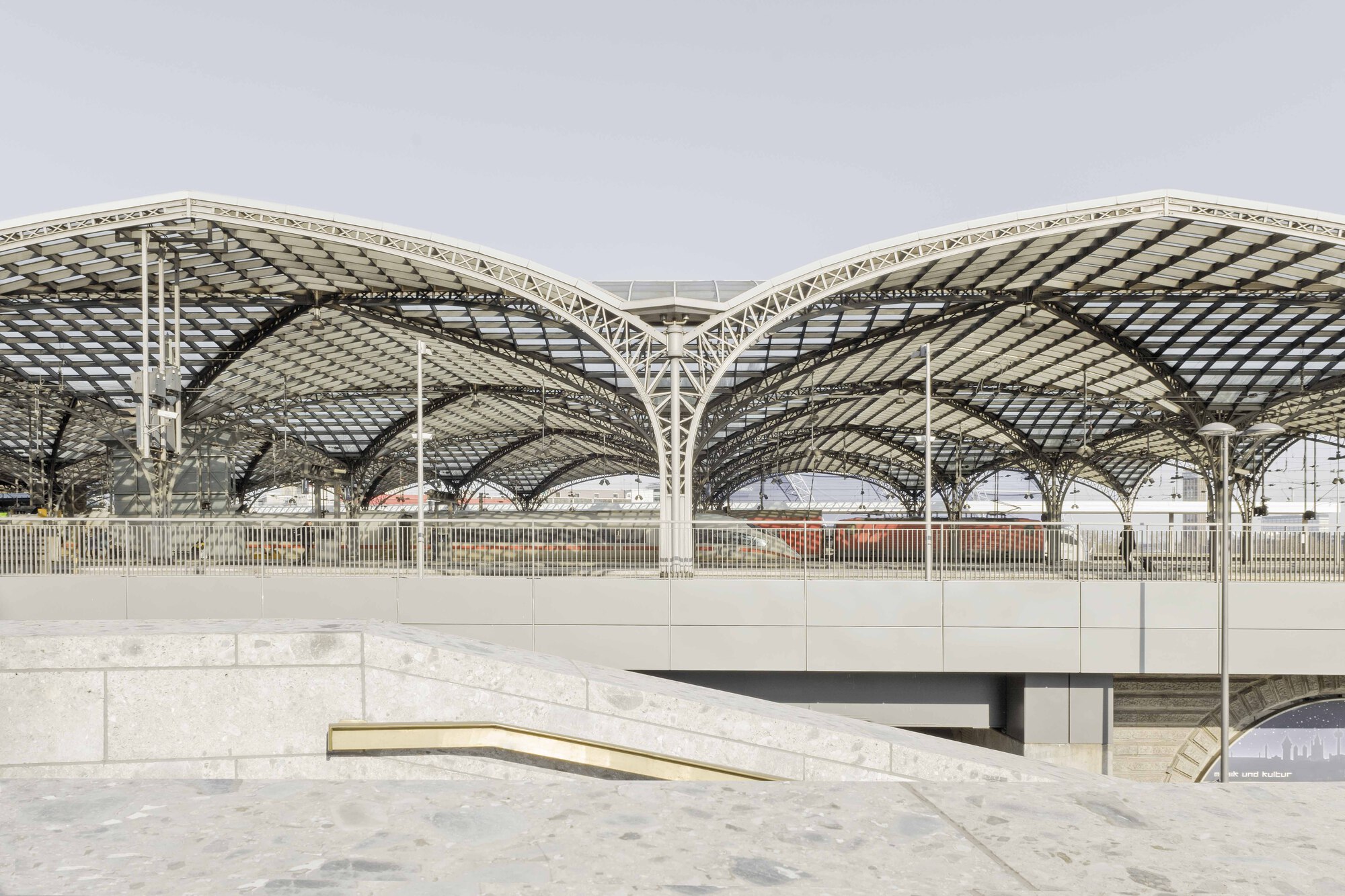 Arched lattice roof structure spanning above a terraced concrete plaza in diffused daylight