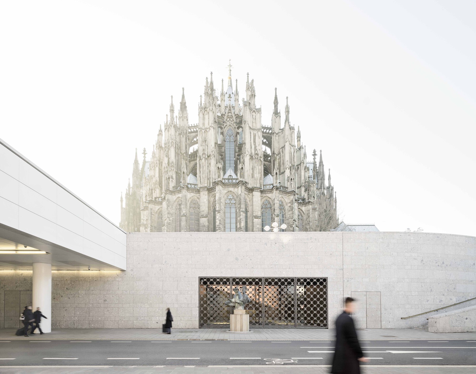 Low stone facade with patterned metal screen framing a courtyard beneath a gothic cathedral on an overcast day
