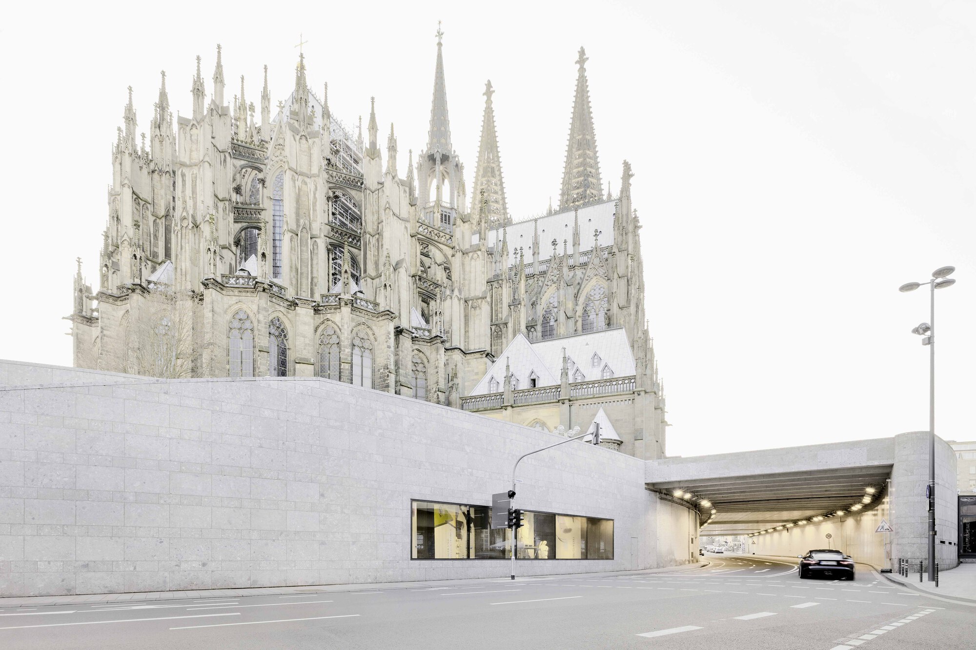 Illuminated tunnel entrance below limestone walls with a gothic cathedral rising behind under grey skies