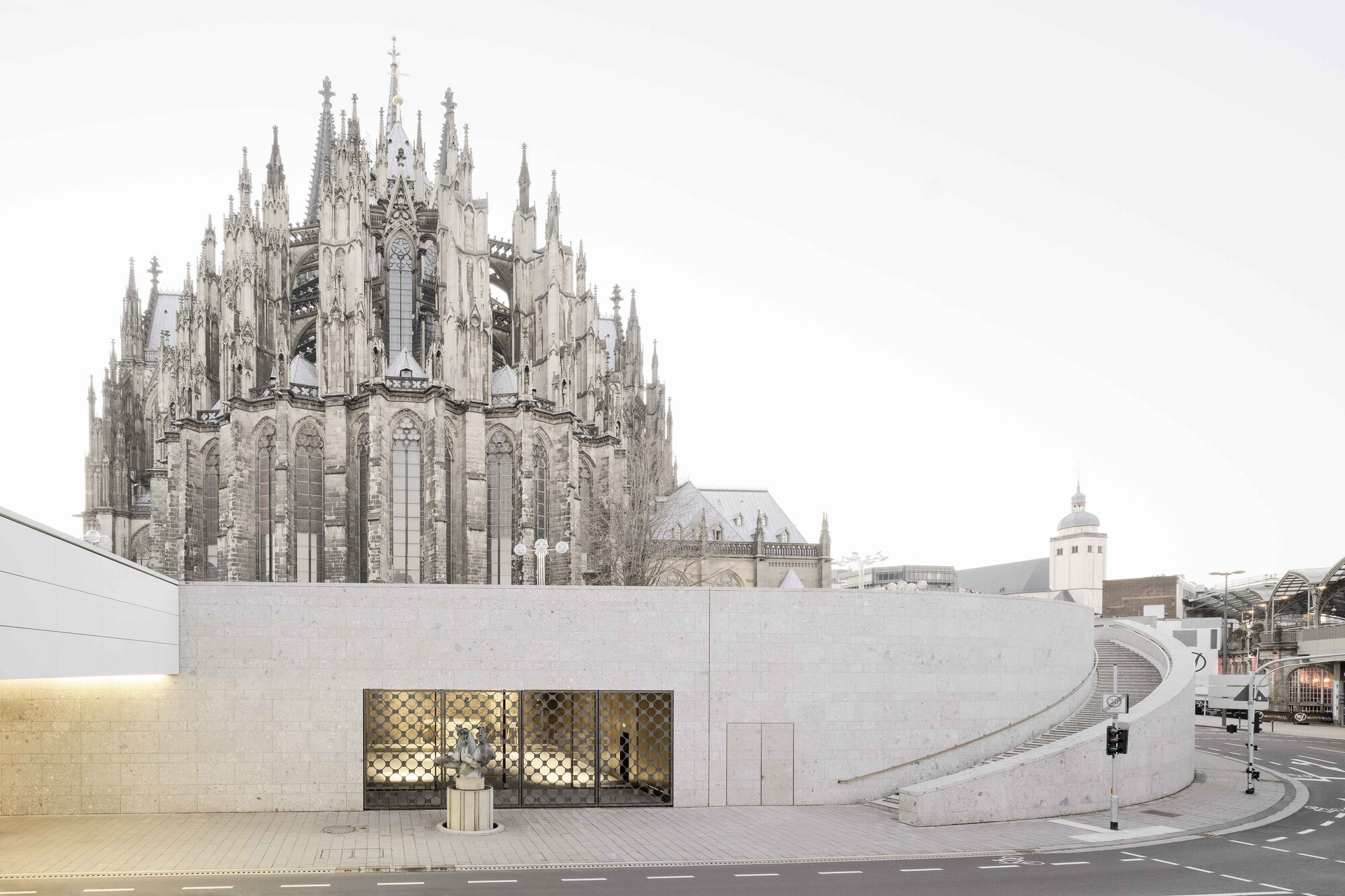 Street view of stone facade with patterned screen windows beneath a gothic cathedral and overcast sky