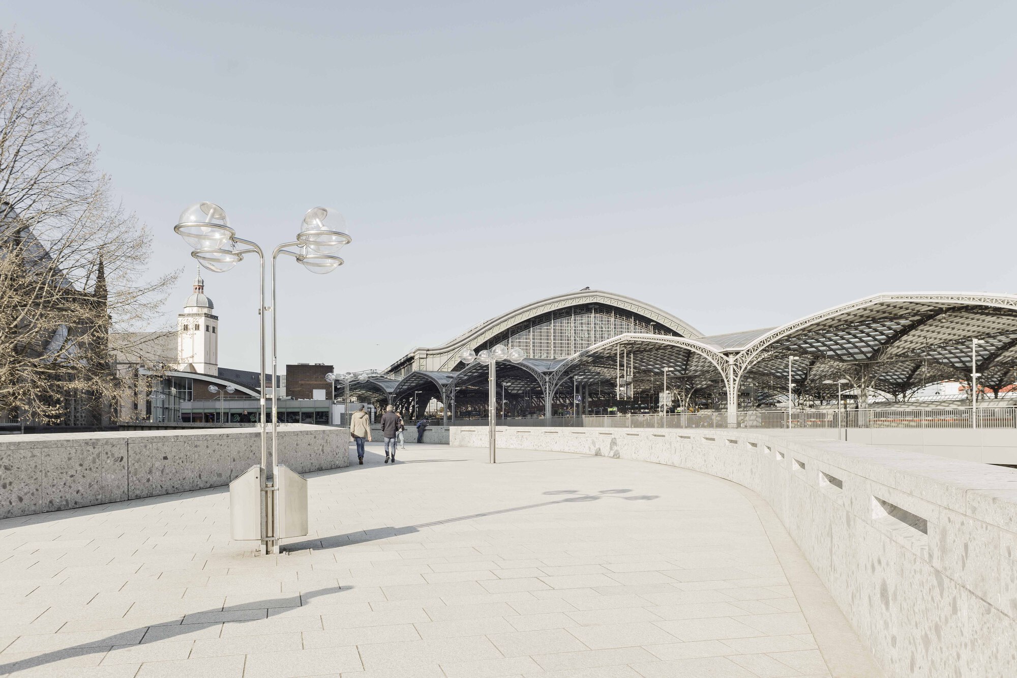 Curved pedestrian plaza with granite pavers leading toward the vaulted station canopies under an overcast sky