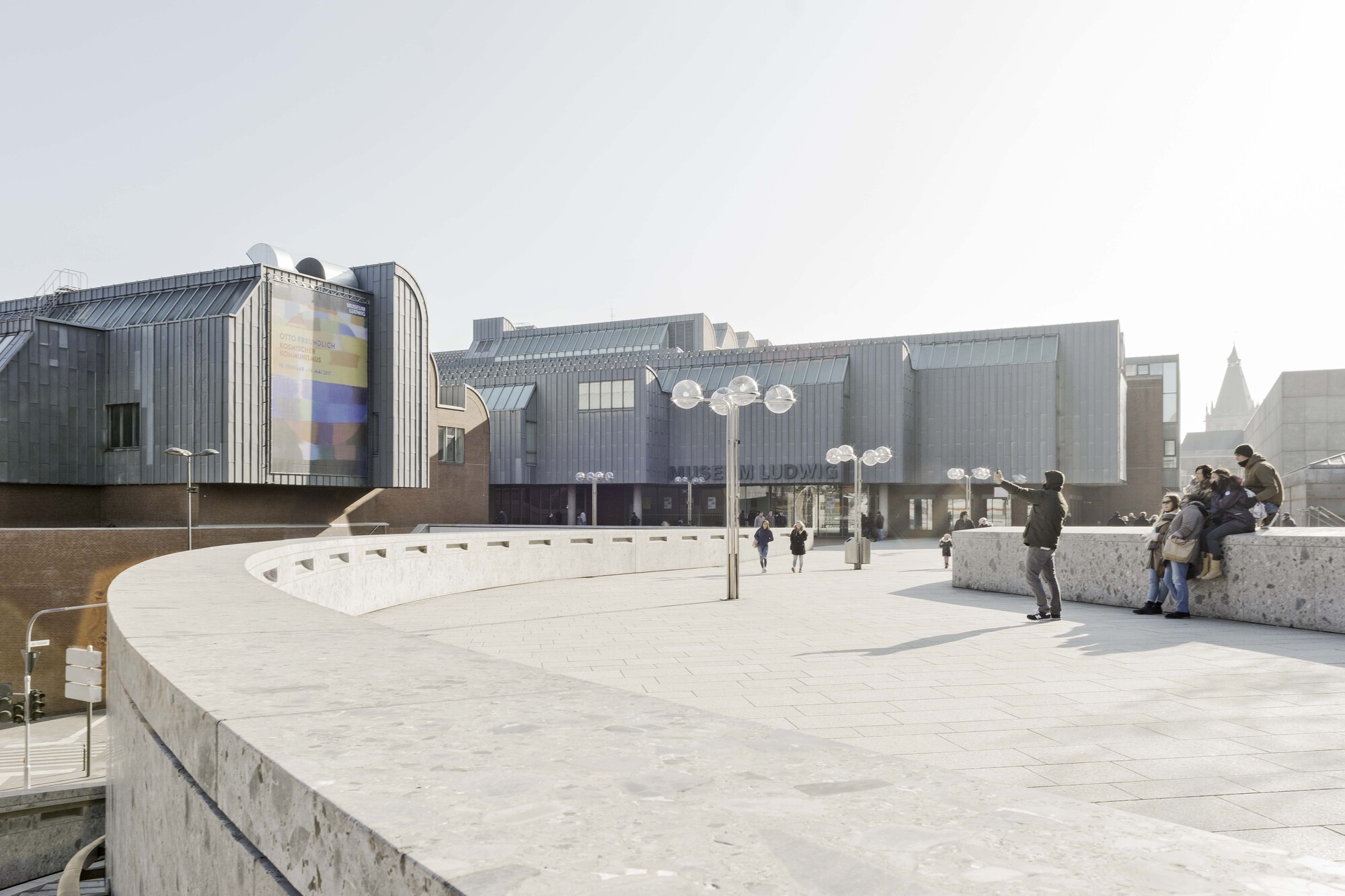 Elevated plaza with concrete benches and paving where visitors gather near the corrugated metal facade