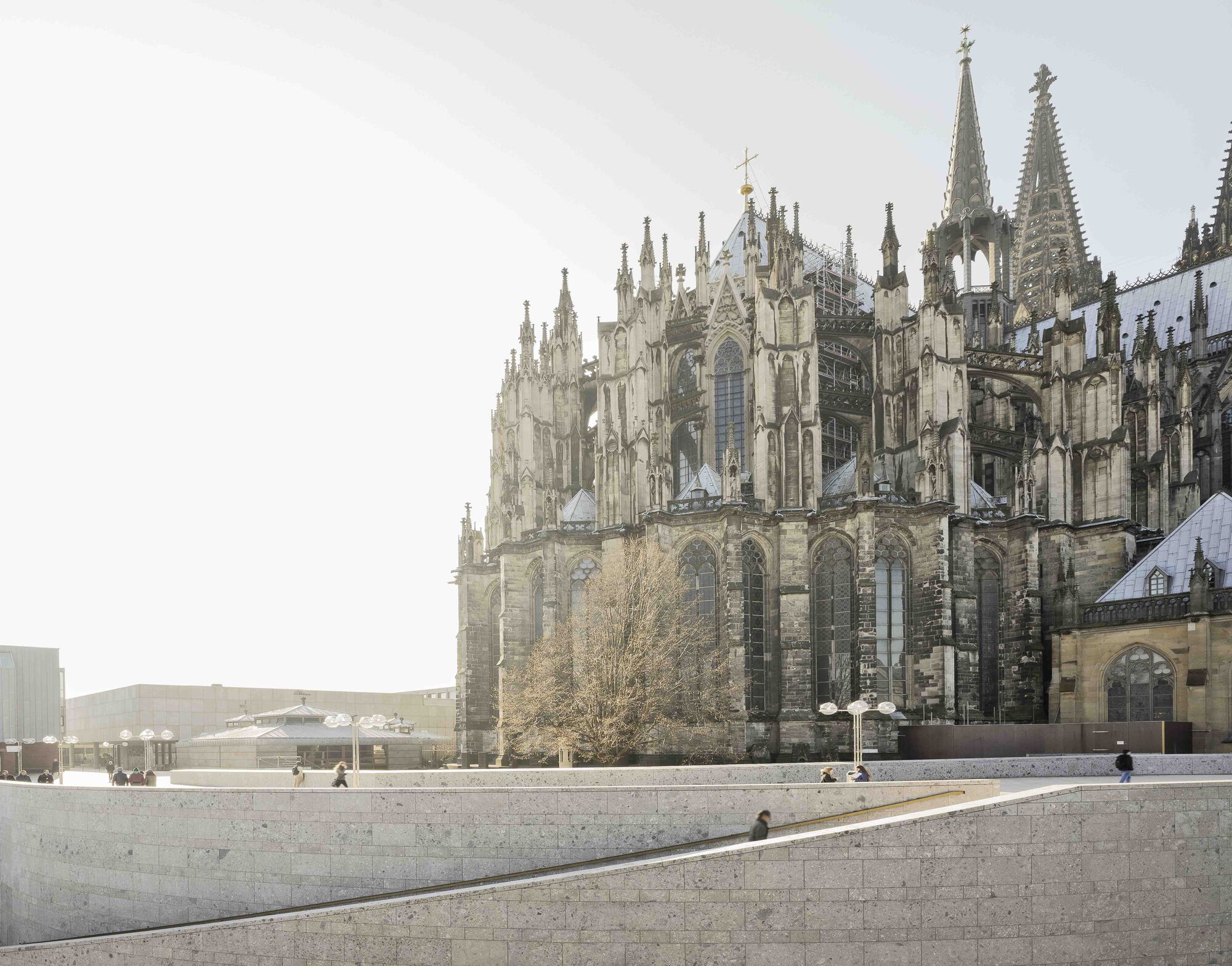 Wide stone plaza with scattered visitors beside the cathedral apse and bare tree in daylight