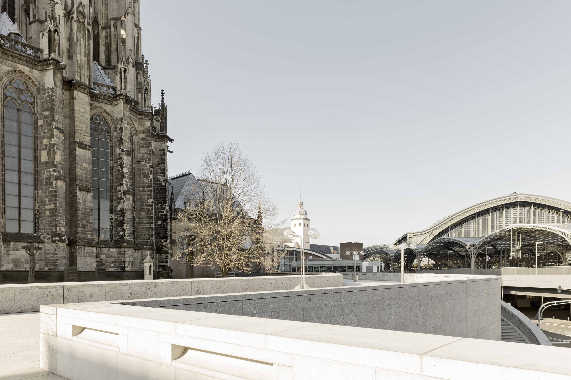 Terraced concrete plaza with linear benches framed by a gothic cathedral and arched train shed
